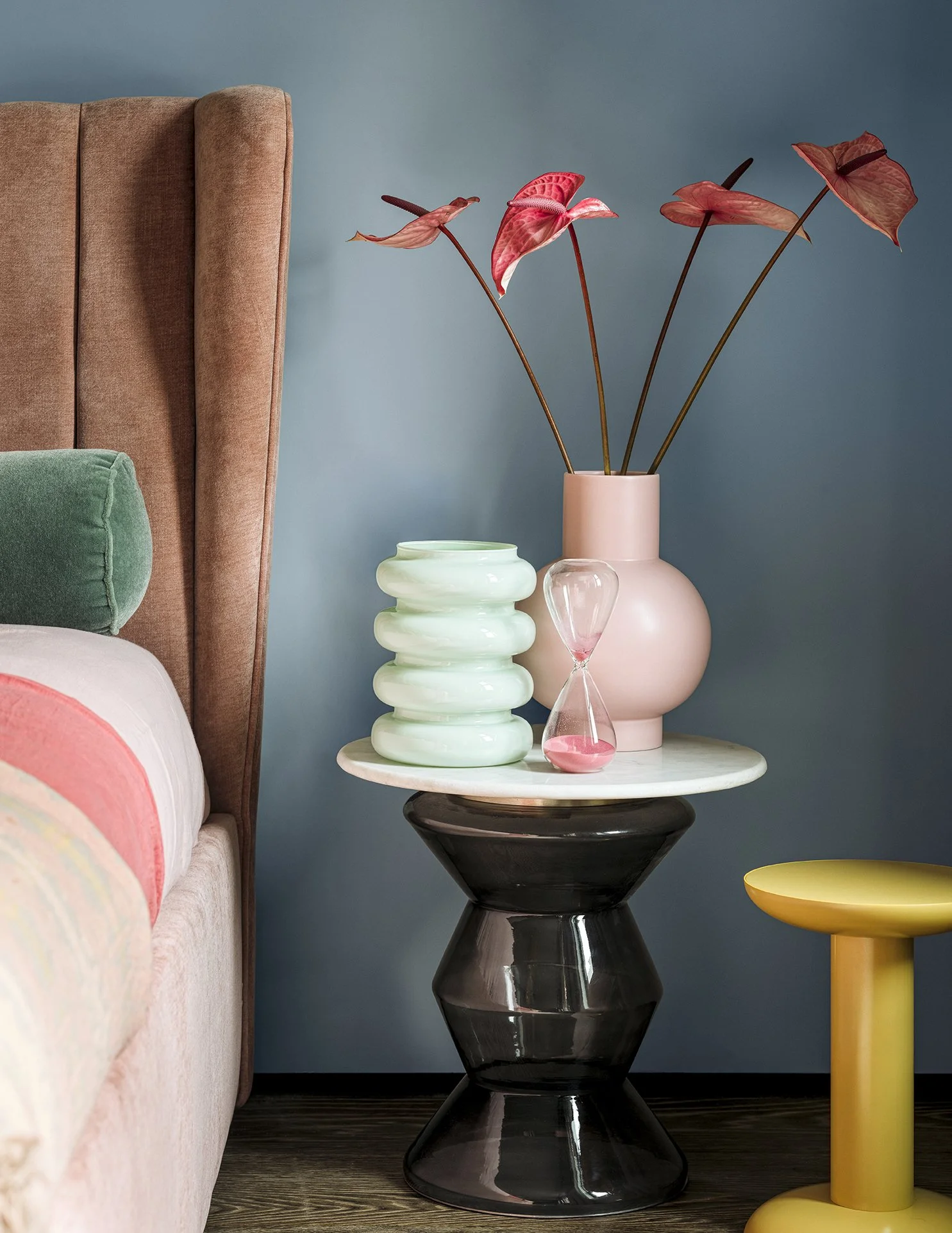 Interior living room decor featuring a side table with decorative vases, a pink vase with red anthurium flowers, and a layered green and white vase. A glimpse of a bed with a pink and white blanket and a pink headboard.