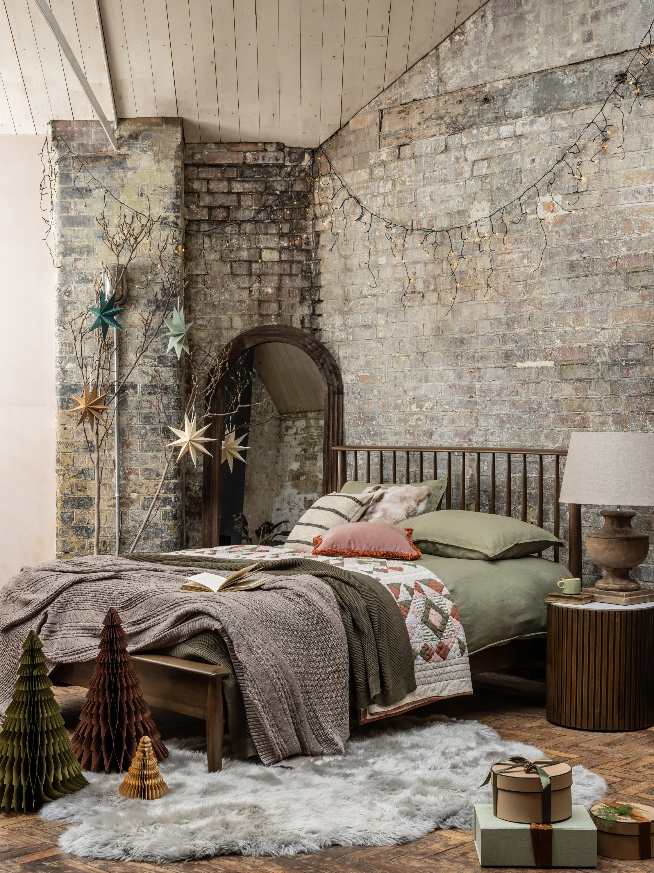 A cozy bedroom with a bed covered in green and patterned bedding, decorated with pillows, on a fluffy white rug. Christmas trees and star ornaments are placed nearby, and string lights hang from the exposed brick and wooden ceiling.