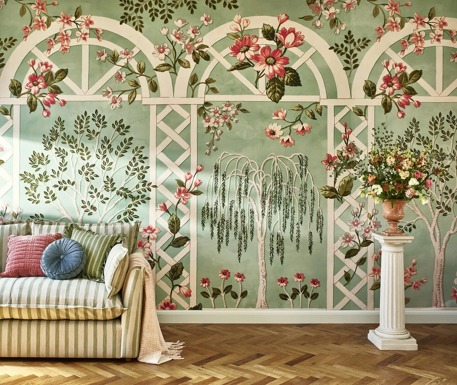Decorative room wall with floral and trellis mural, striped sofa with pillows, and a floral arrangement on a pedestal