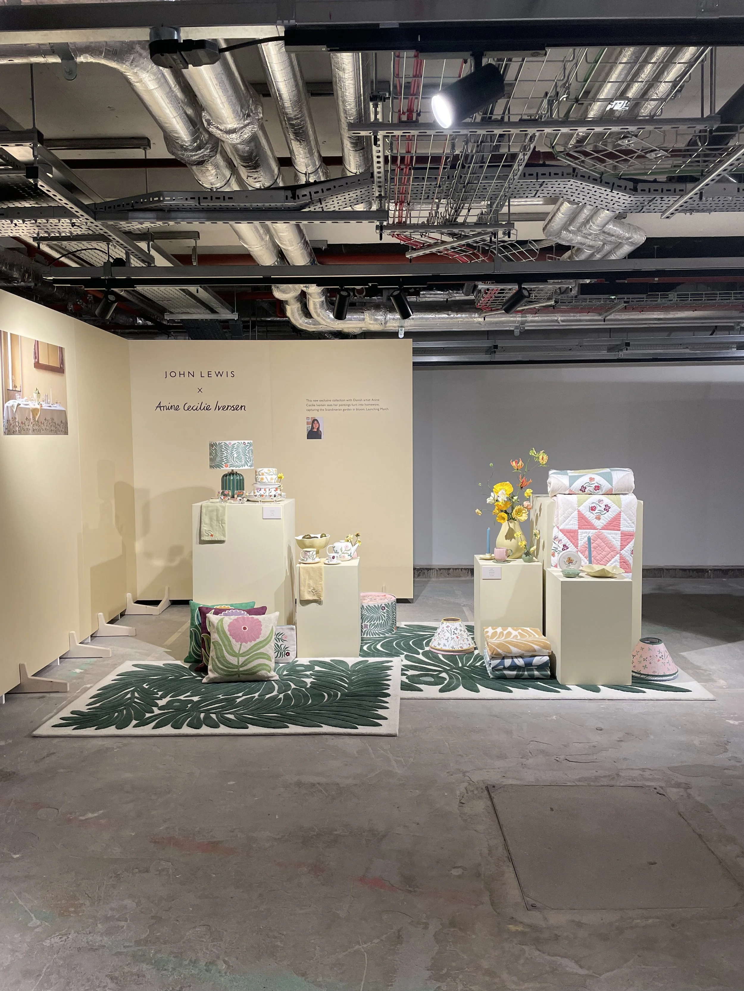An art installation titled 'John Lewis x Anne Cecile Iversen' featuring home decor items such as colorful pillows, lampshades, vases with flowers, and ceramic dishes, arranged on a green tropical-patterned rug and white pedestals in a gallery setting