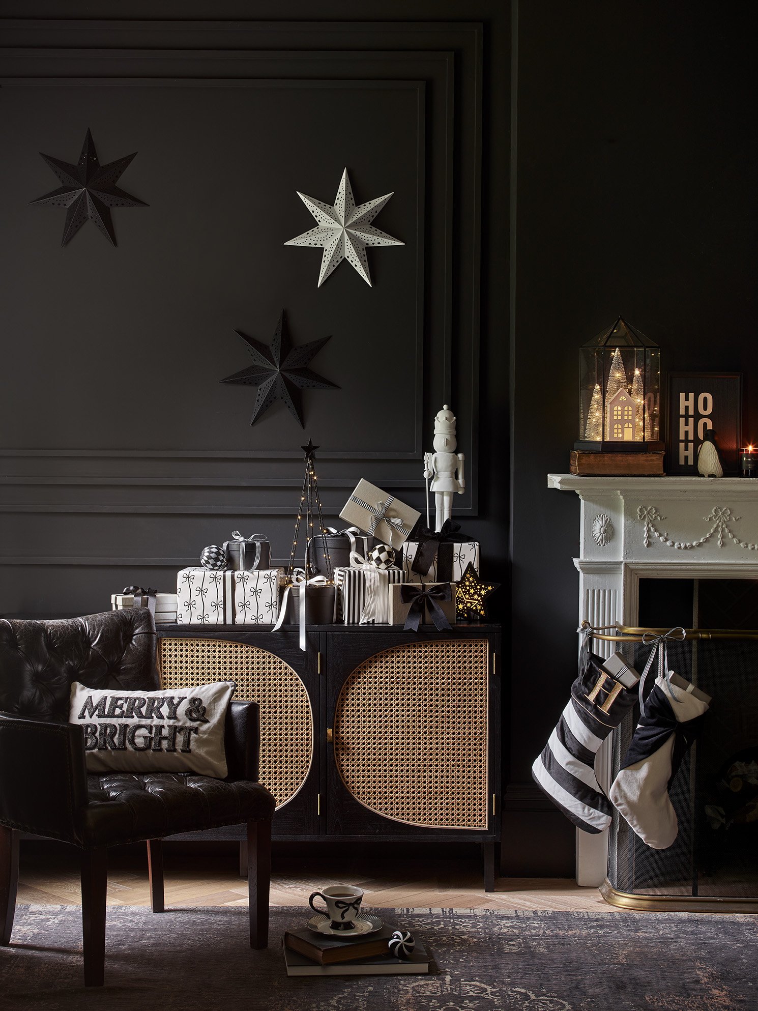 A cozy holiday living room corner with black walls and Christmas decorations, including black and white presents, stockings, and star ornaments on the wall, a white decorative fireplace with a house-shaped lantern, and a black armchair with a pillow 