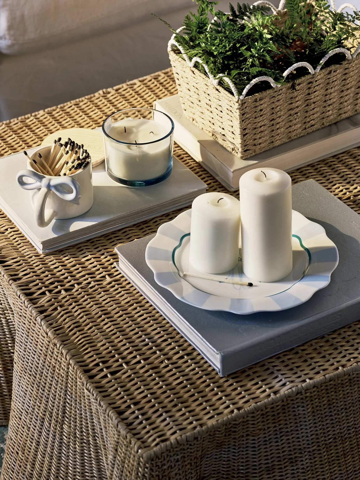 Decorative table setup on a rattan table with candles, books, a small plant arrangement, and a container of matches.