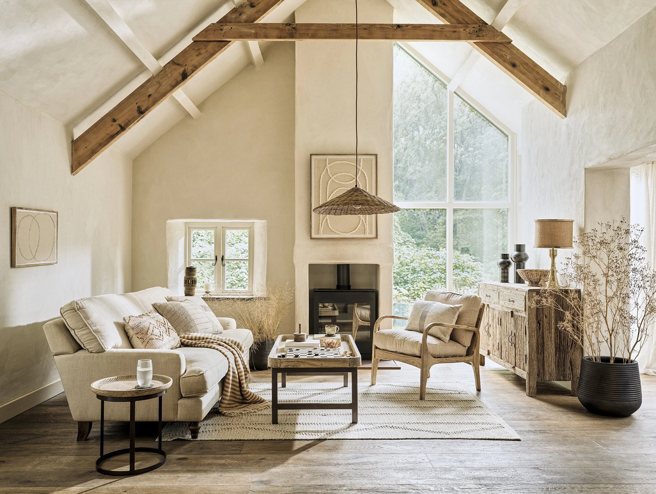 A cozy living room with vaulted ceiling and exposed wooden beams, large triangular and rectangular windows, a white sofa with pillows, a wooden armchair, a sideboard, a fireplace, and decorative lighting with natural light streaming in.