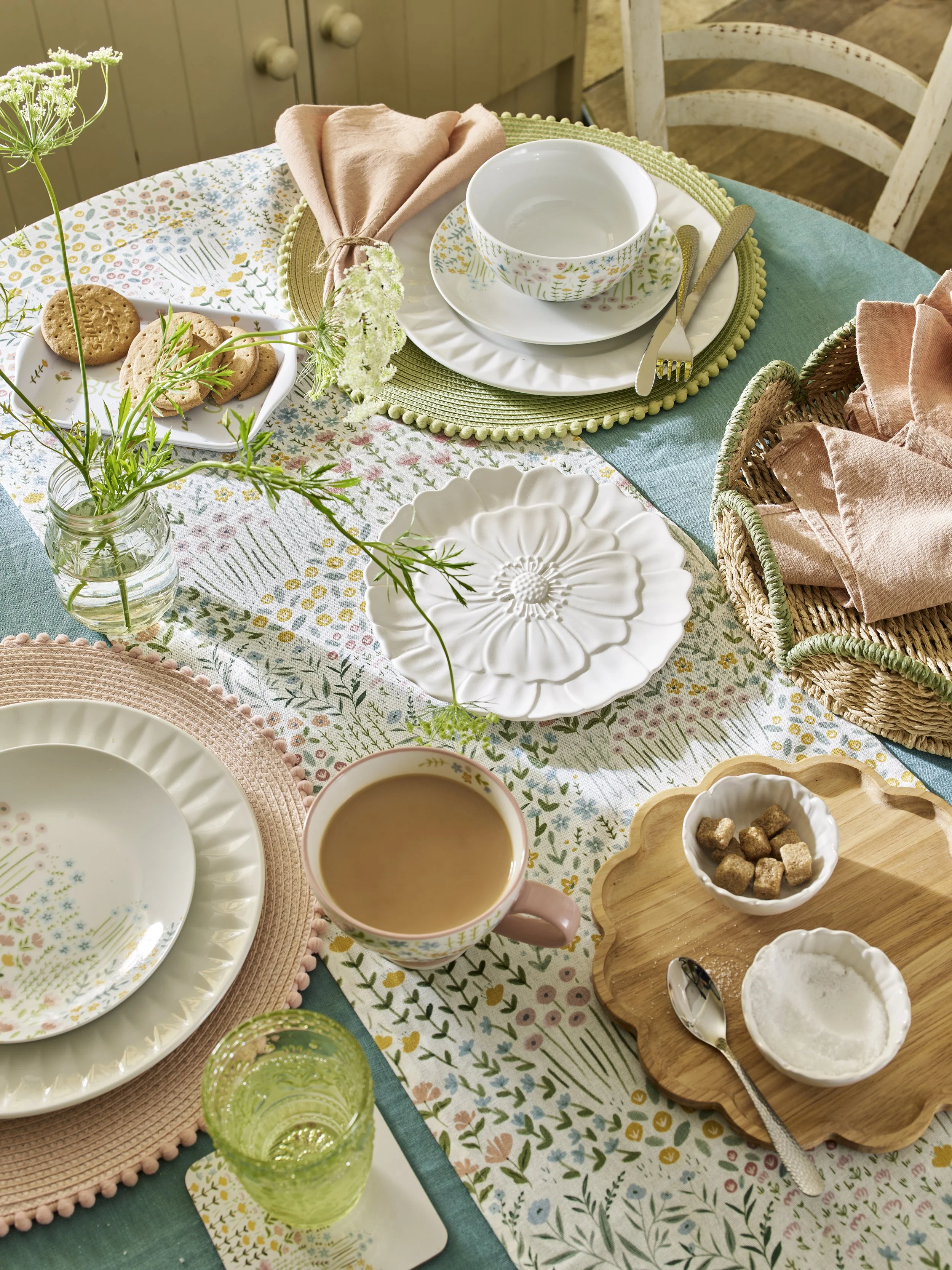 A table set for a meal with floral-patterned plates, a cup of coffee, a glass of water, cookies, sugar cubes, and a flower in a small vase on a colorful tablecloth.