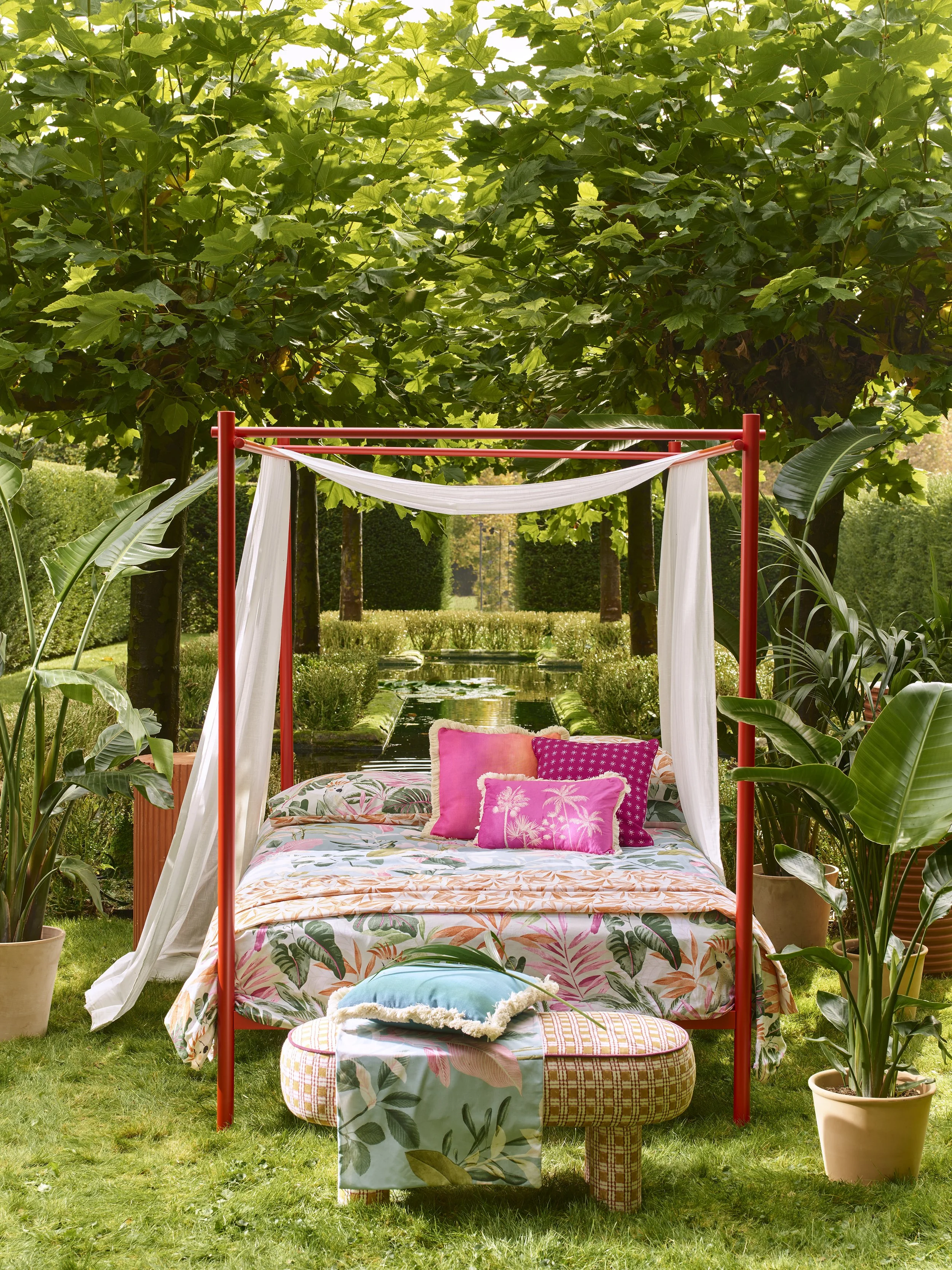 Outdoor canopy bed with white drapes, pink and patterned pillows, floral bedding, and a bench with pillows, surrounded by lush green plants and trees in a garden.