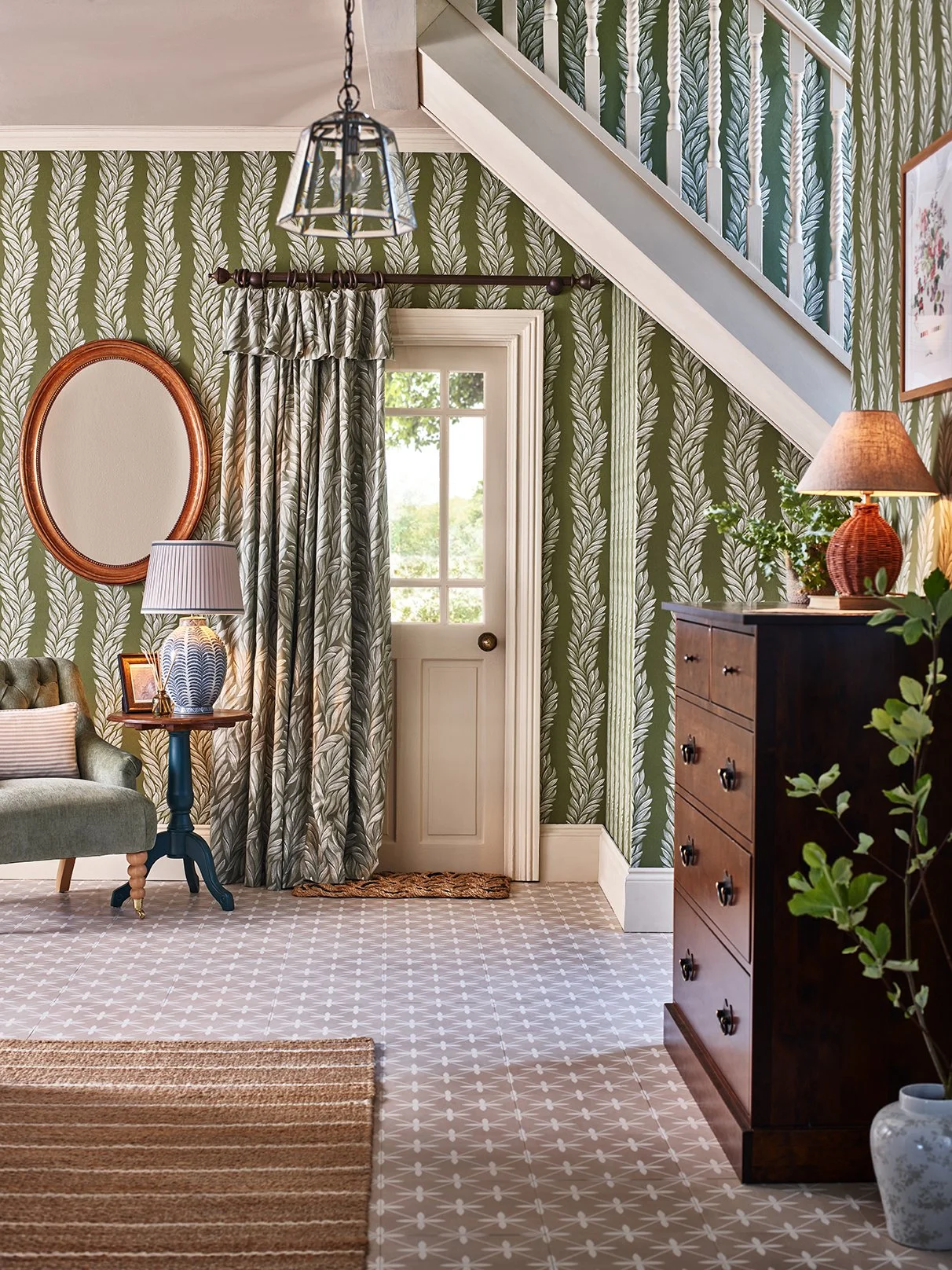 Interior view of a room with green leaf-patterned wallpaper, a window with curtains, a door, a wooden dresser, a small side table with a lamp, a mirror, and a sofa.