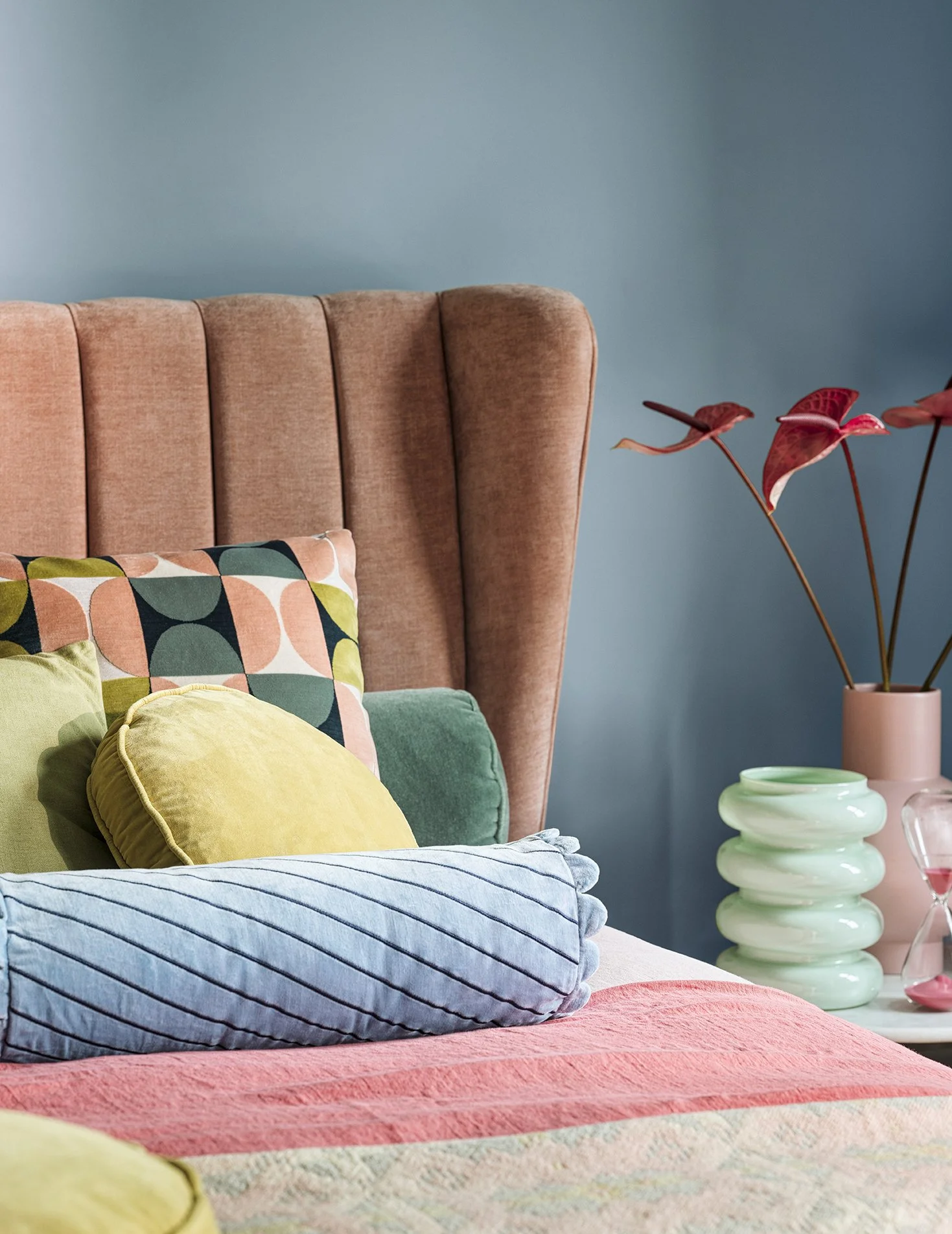 A bedroom corner with a pink upholstered headboard, colorful pillows, and a pastel-colored bedside table with decorative items and a potted plant.