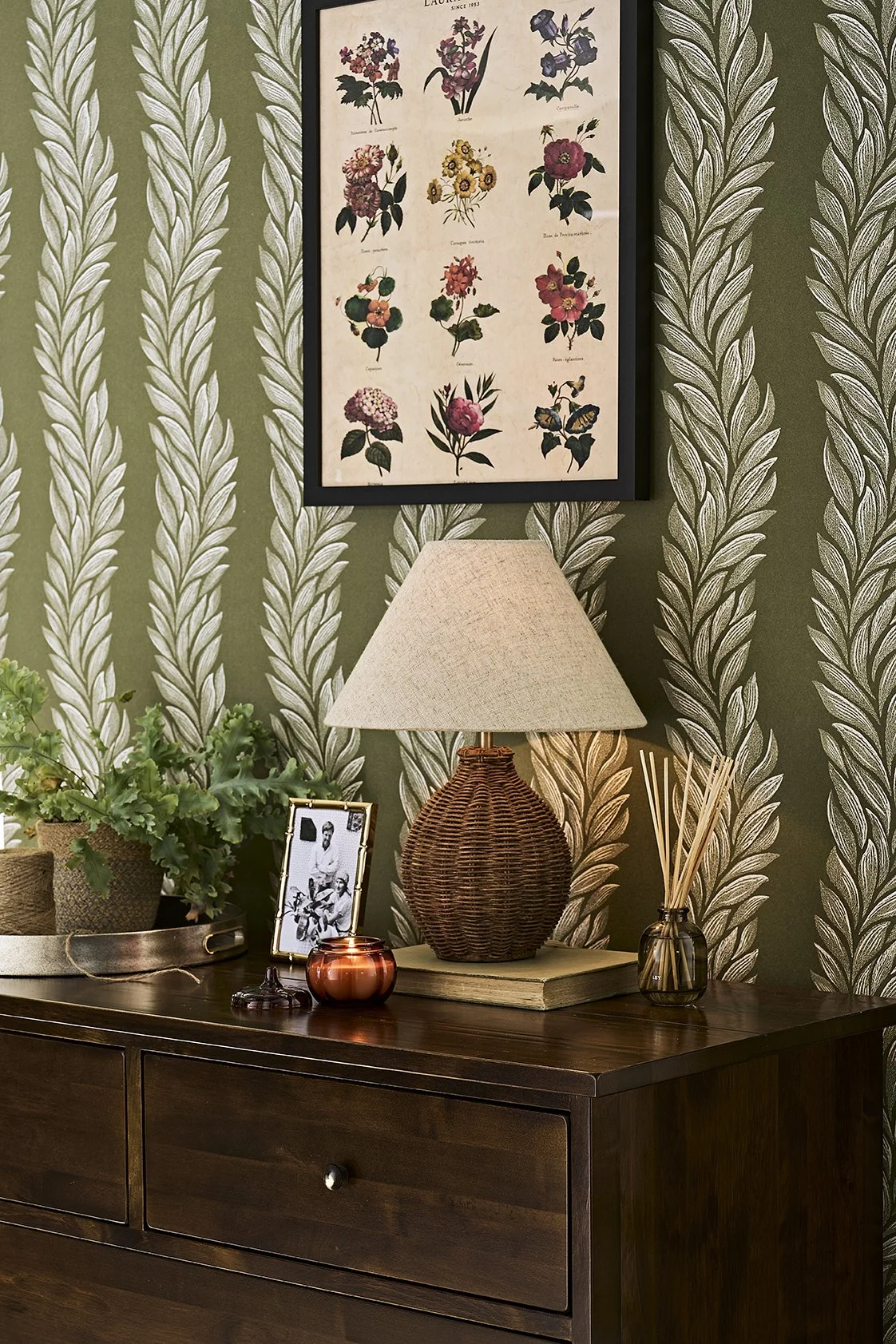 Decorative interior scene with a dark wooden dresser against a green, leafy patterned wallpaper. On the dresser, there is a potted plant, a framed black and white photo, a small copper-colored candle holder, a large wicker table lamp with a beige lam