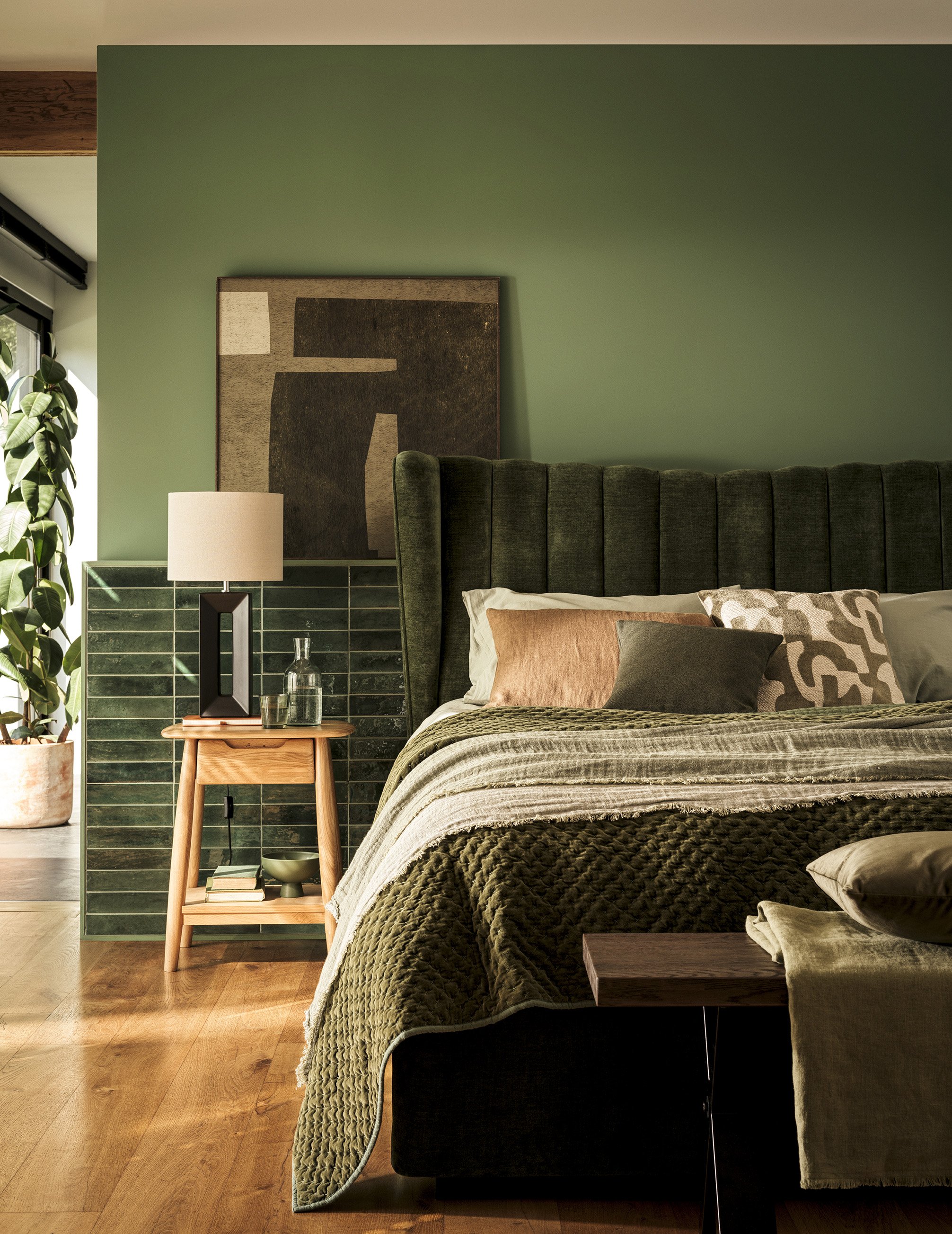 Contemporary bedroom with green upholstered headboard, abstract artwork, side table with lamp, green tiled wall, glass bottle, and pillows