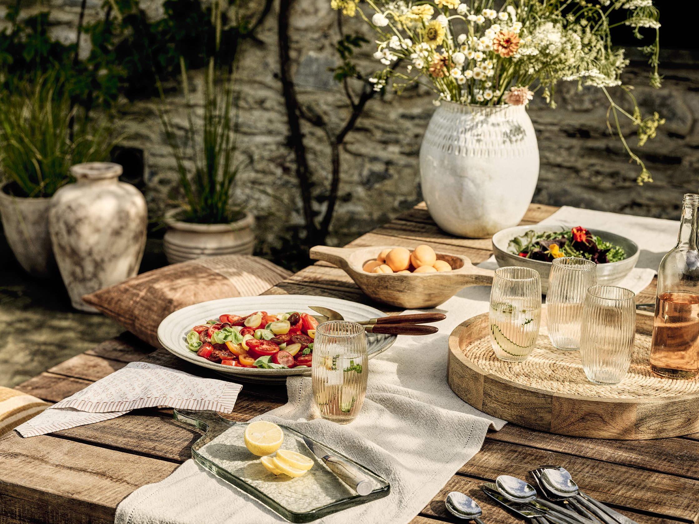 Outdoor dining table set with a variety of salads, lemon slices, and drinks, decorated with flowers and plants, in a rustic garden setting.