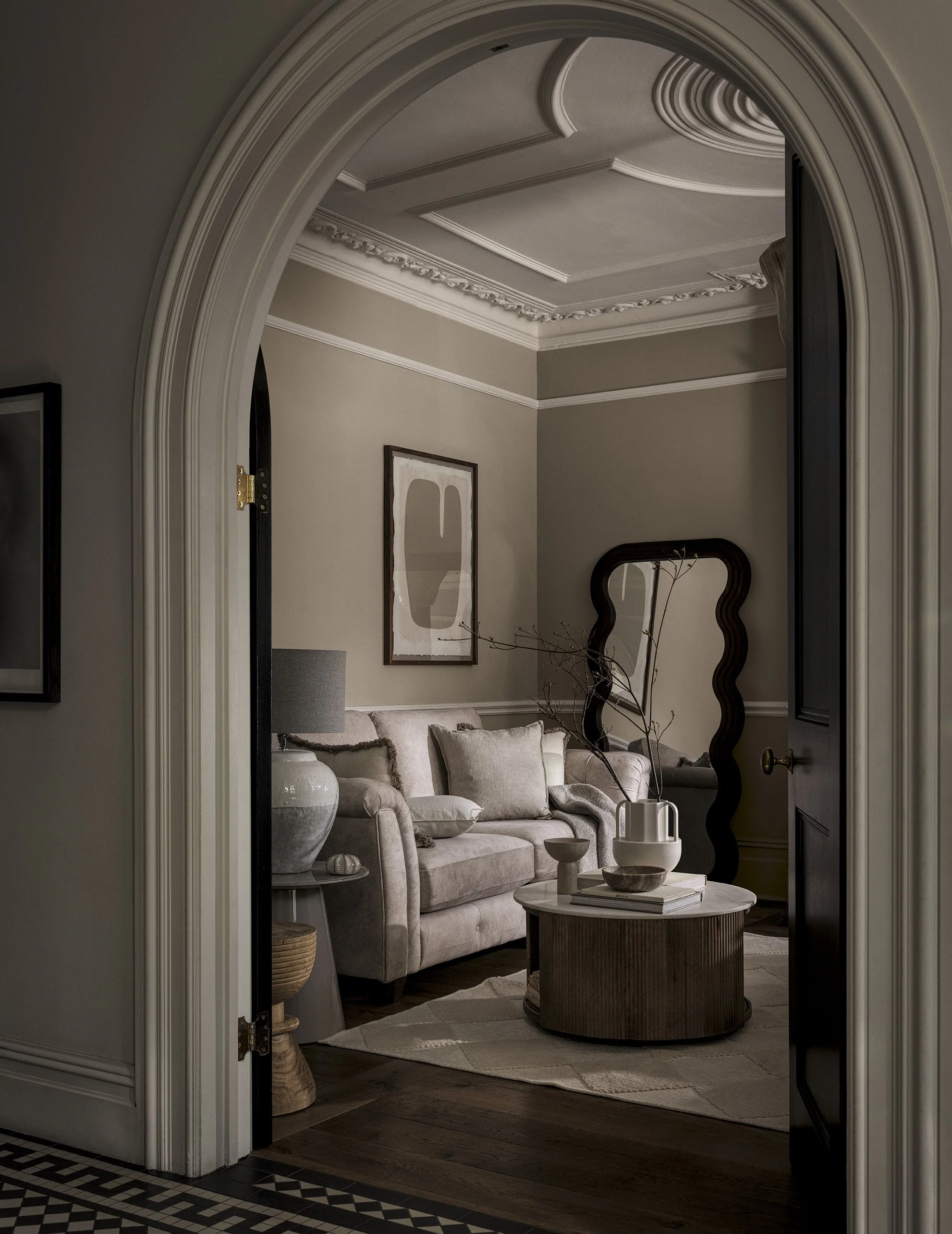 Living room with beige sofa, dark wood coffee table, modern black mirror, abstract art on wall, decorative pillows, and a grey lamp on side table, seen through an arched doorway.