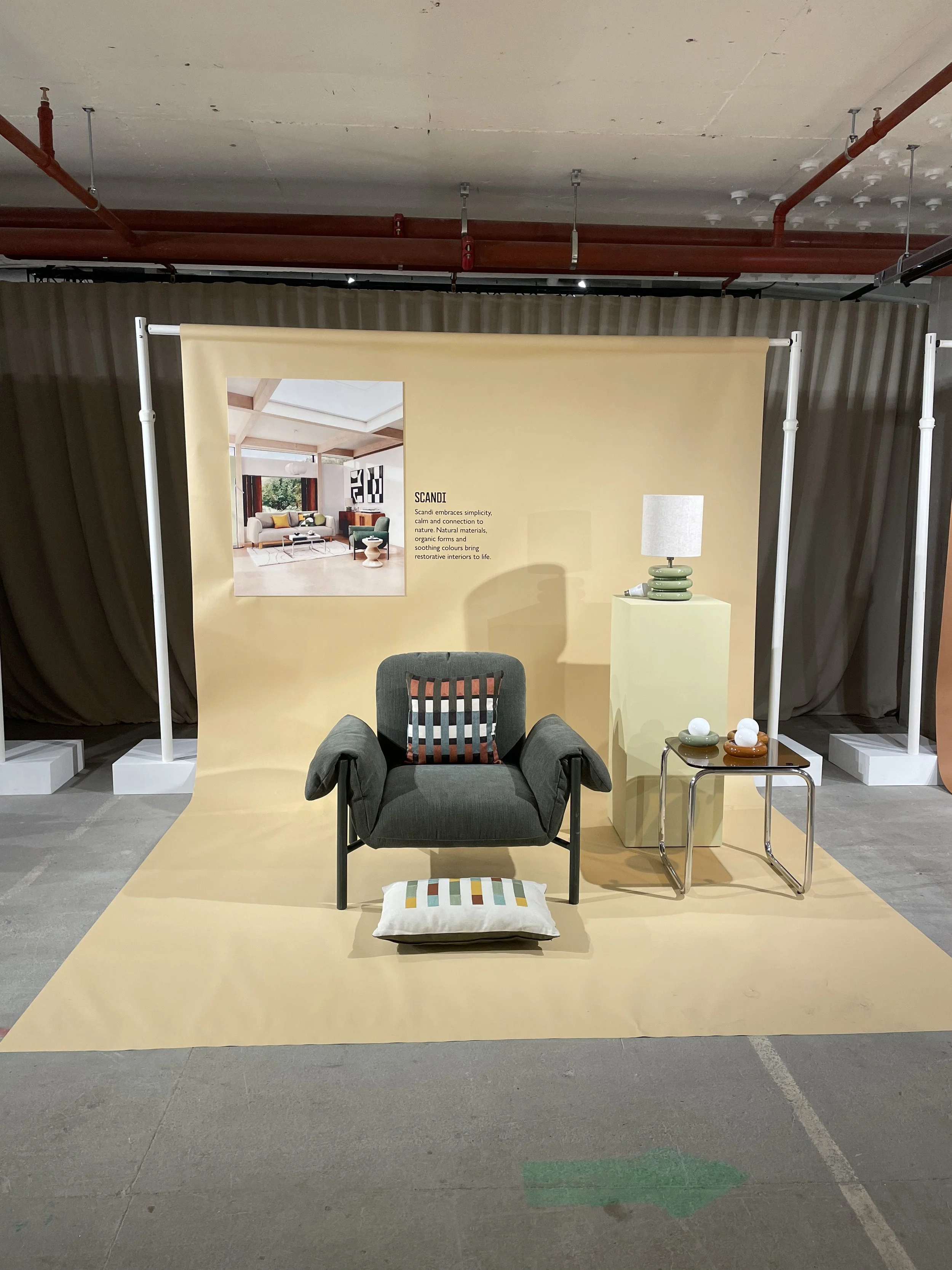 A lounge chair with a cushion in front, a small side table with decorative vases, a lamp, and a yellow wall in the background displaying a room interior photo and description.