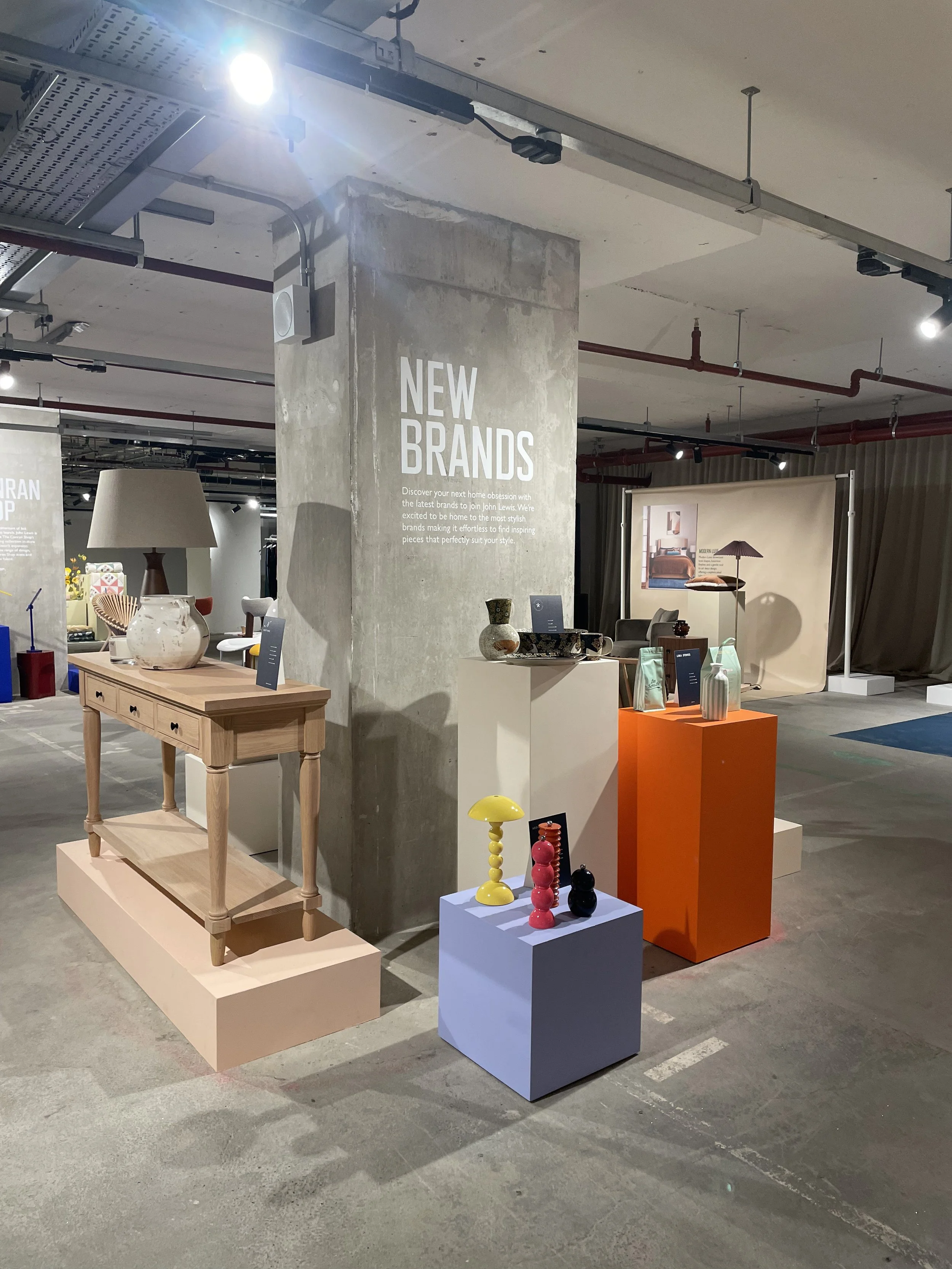 Display of new home decor and furniture items in a showroom with a concrete pillar, tables, and colorful decorative objects.