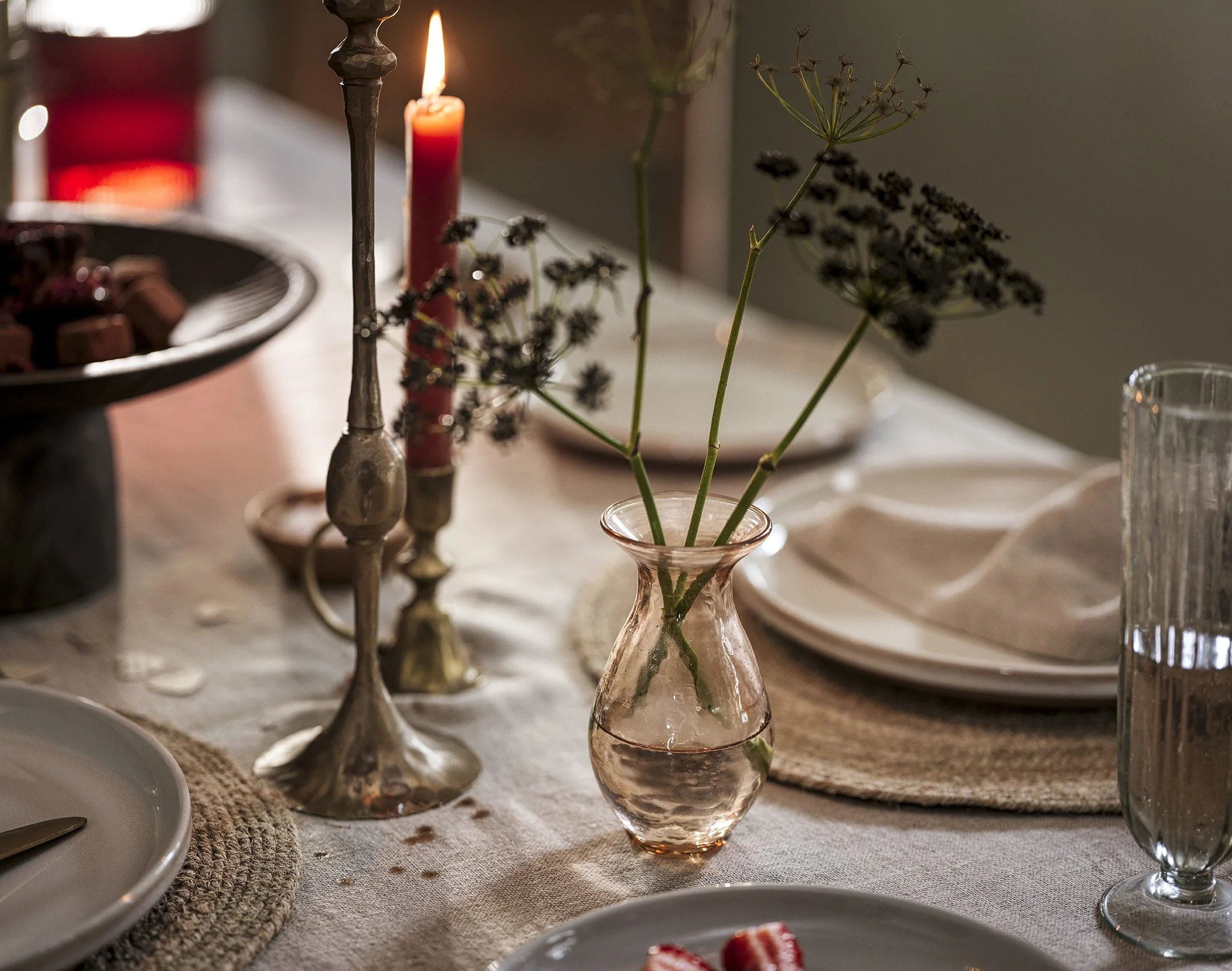 A table setting with a pink glass vase holding greenery, a lit red candle in candle holder, plates, glasses, and a bowl of chocolates in the background, creating a cozy and elegant atmosphere.