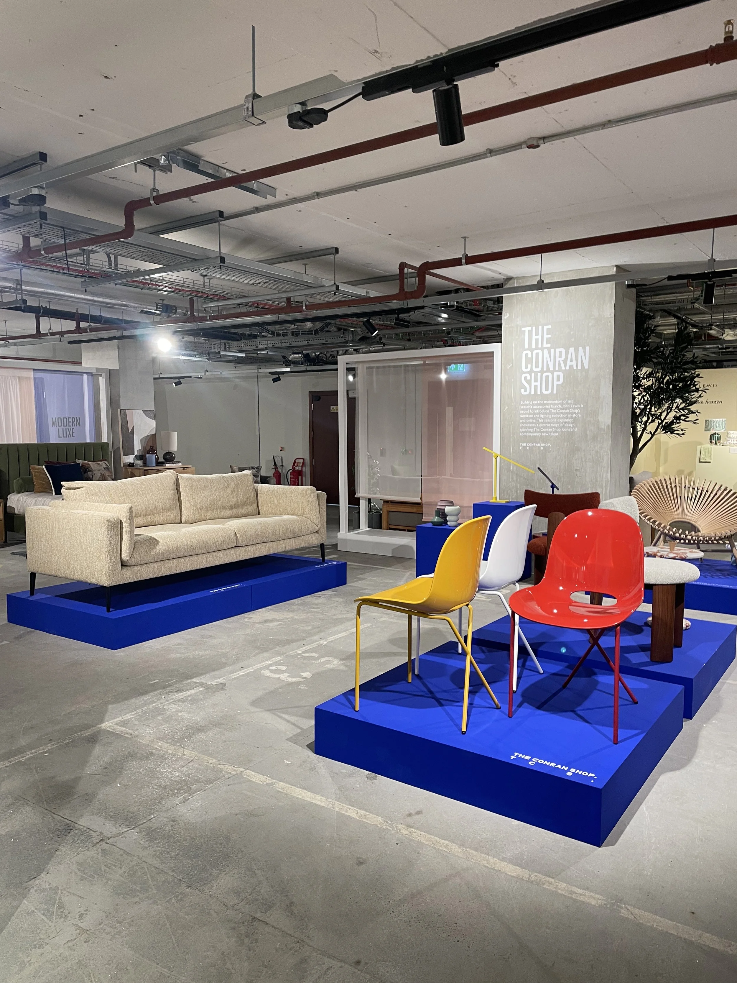 Display of various colorful chairs and furniture pieces at The Conran Shop, including a beige sofa, yellow, white, and red chairs on blue platforms, in an industrial-style interior.