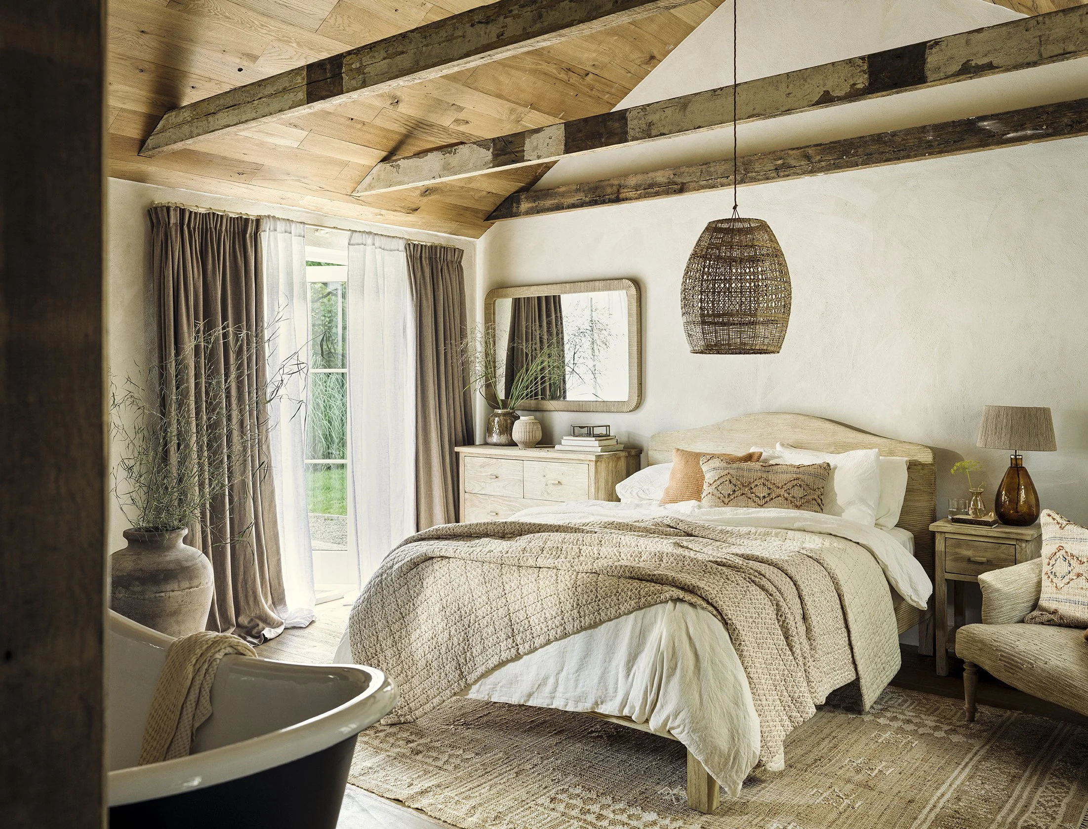 A cozy bedroom with a neutral color palette, featuring a bed with beige bedding, wooden furniture, a large window with brown and white curtains, a wicker pendant light, a mirror, a lamp, and decorative pillows, with a bathtub partially visible in the