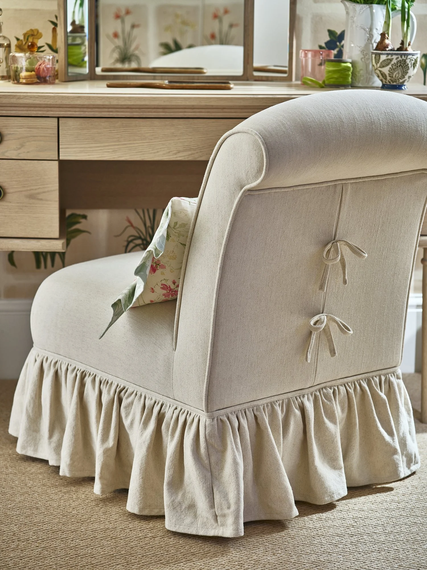 A cream-colored upholstered armchair with a ruffled skirt, side bows, and a floral pillow in front of a wooden desk with a mirror and various small decorative plants and items.