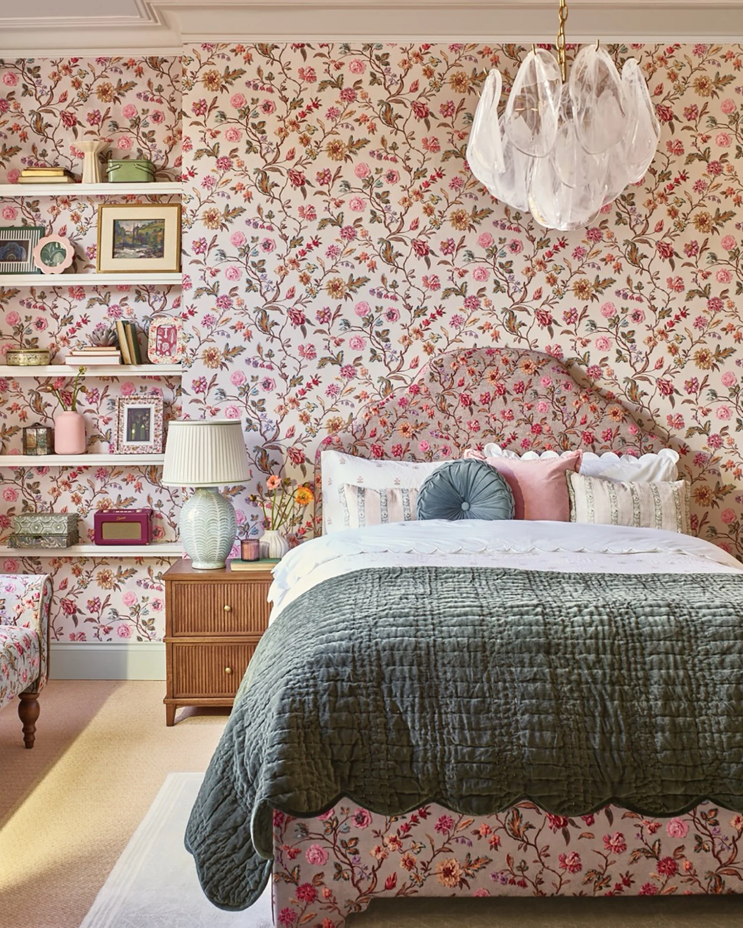 A cozy bedroom with floral wallpaper, a bed with a floral headboard, multiple decorative pillows, a green quilt, and a white bedspread. A bedside wooden nightstand holds a lamp, flowers, and small decorative items. There are built-in shelves with boo