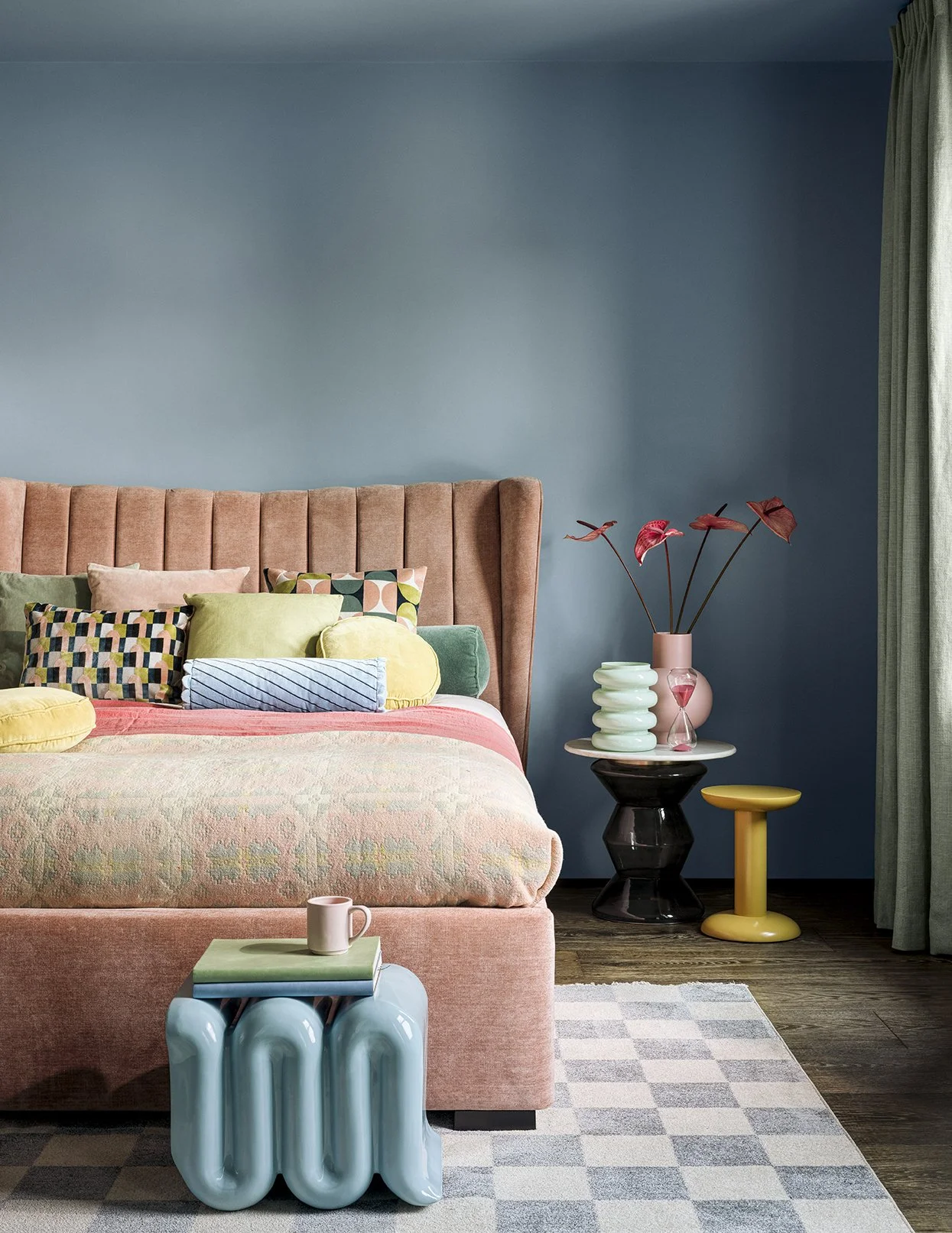 Colorful bedroom with pink upholstered bed, stacked cushions, side table with decorative vases and lamp, curtain, and a patterned area rug.