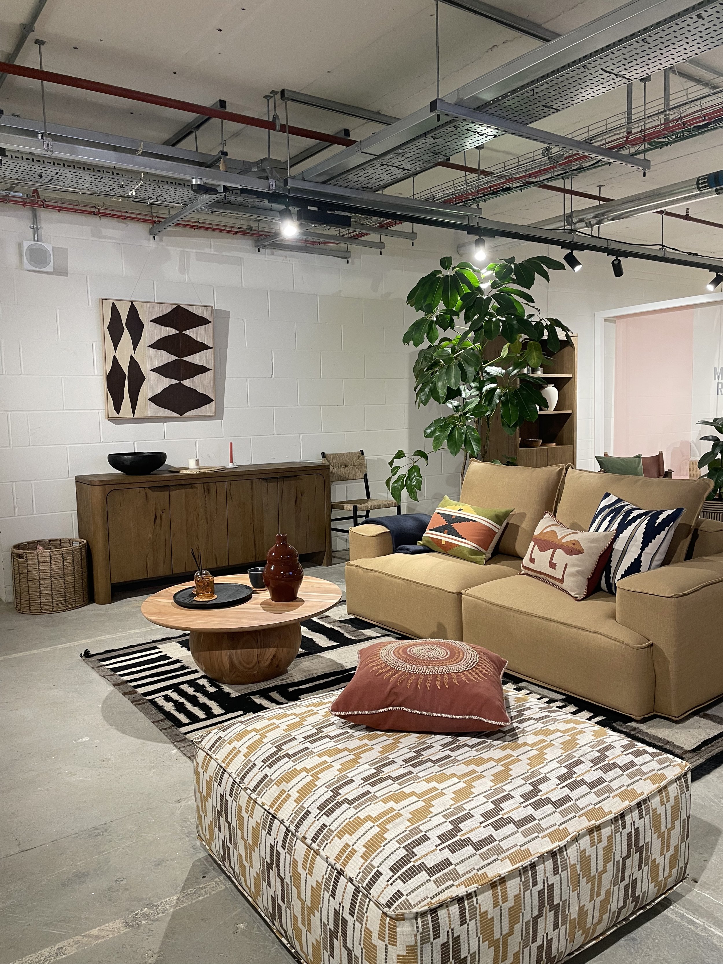 Living room with beige sofa, patterned pillows, round wooden coffee table, coffee table with decorative items, geometric patterned ottoman, large green plant, wooden sideboard, wall art, and cinder block walls with visible ceiling pipes and track lig