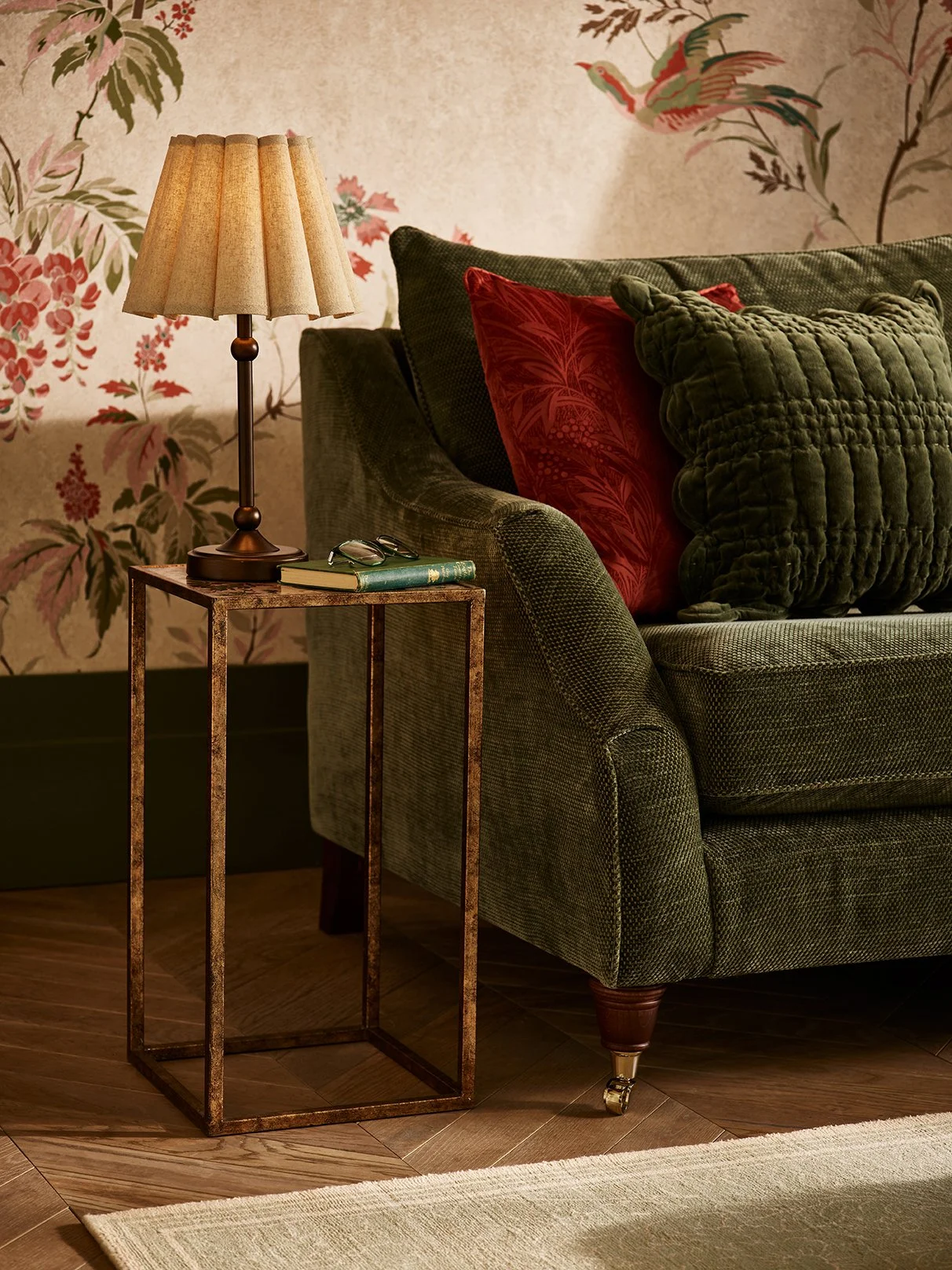 Living room with green sofa, red and green pillows, a side table with a lamp, pair of glasses, and a book, and wallpaper with birds and floral design.