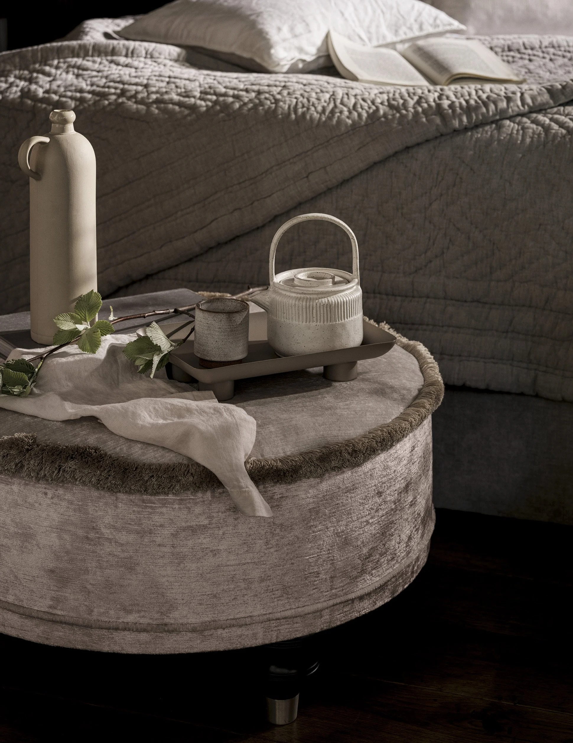 A round ottoman with a textured fabric surface in front of a bed. On the ottoman sits a beige tray holding a white ceramic pitcher, a small dark mug, and a beige teapot. A branch with green leaves and a white cloth are also on the ottoman.