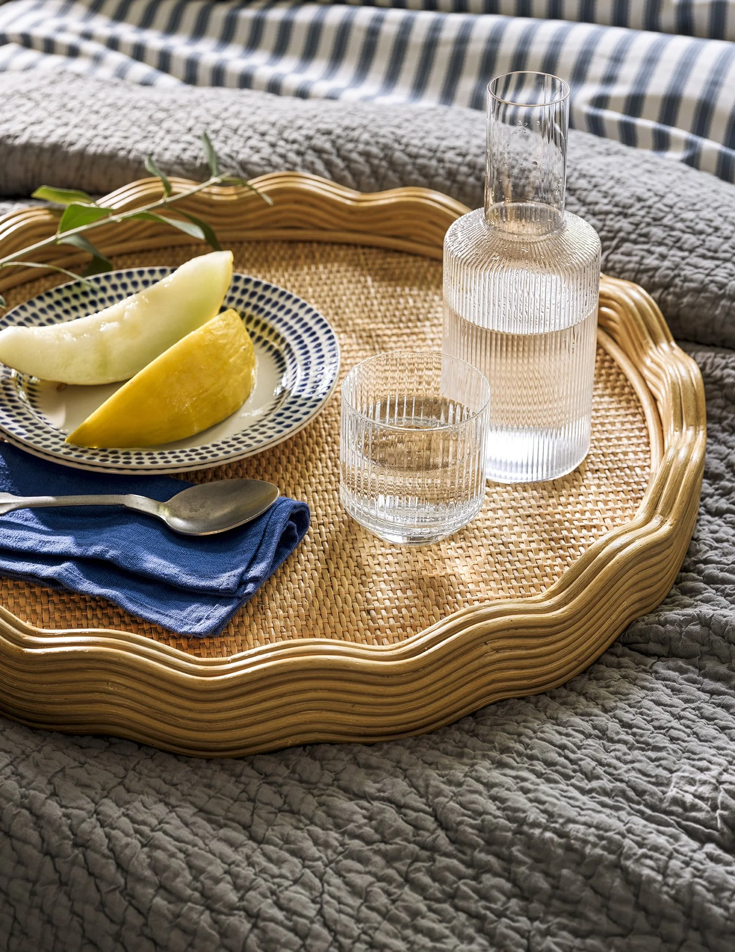 A decorative tray with a glass water decanter, a glass of water, a plate with melon slices, a blue napkin, and a spoon, placed on a quilted bedspread.