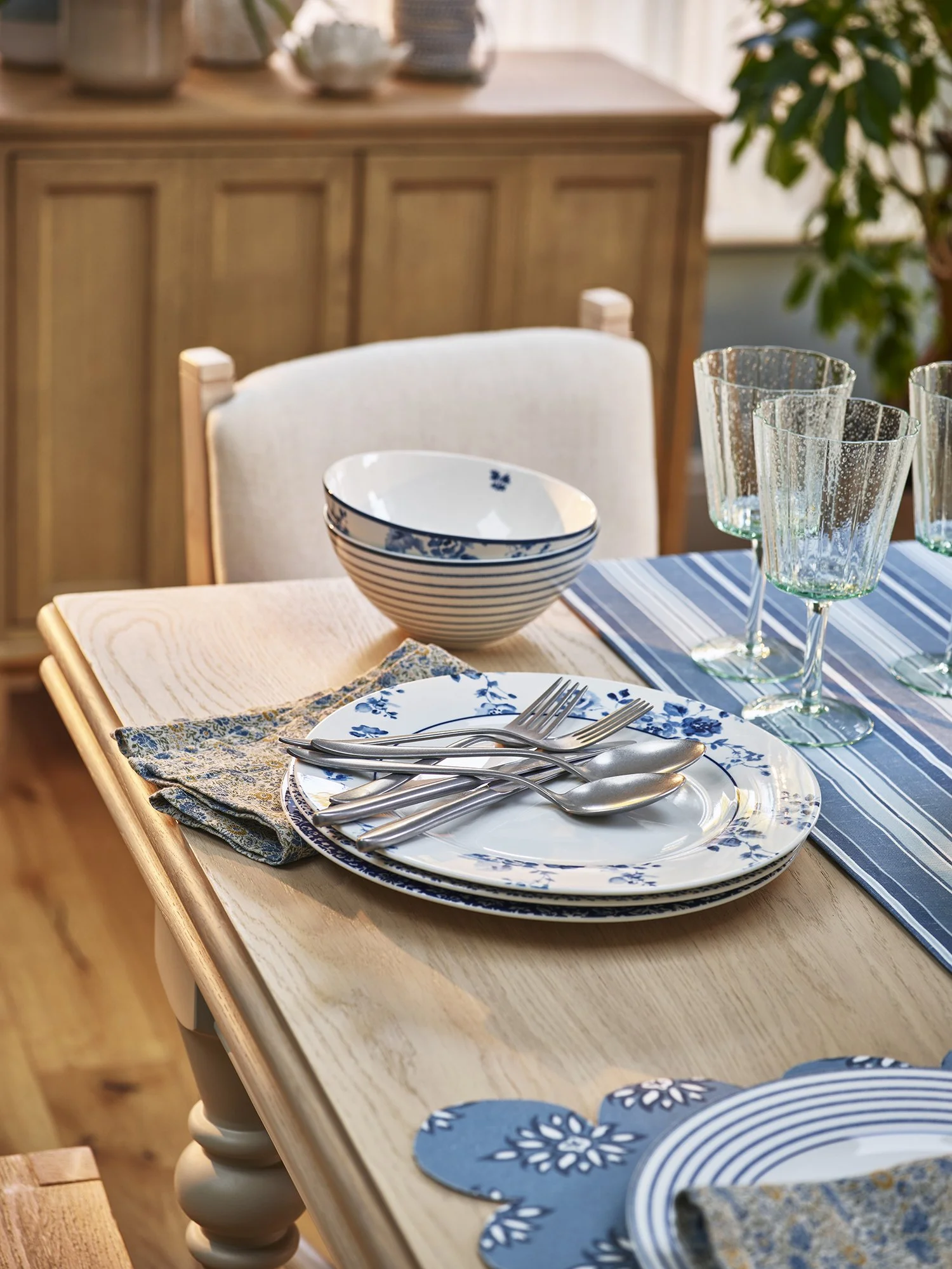 Set dining table with blue and white patterned plates, silverware, clear glasses, and a napkin with a blue floral pattern on a light wooden table.
