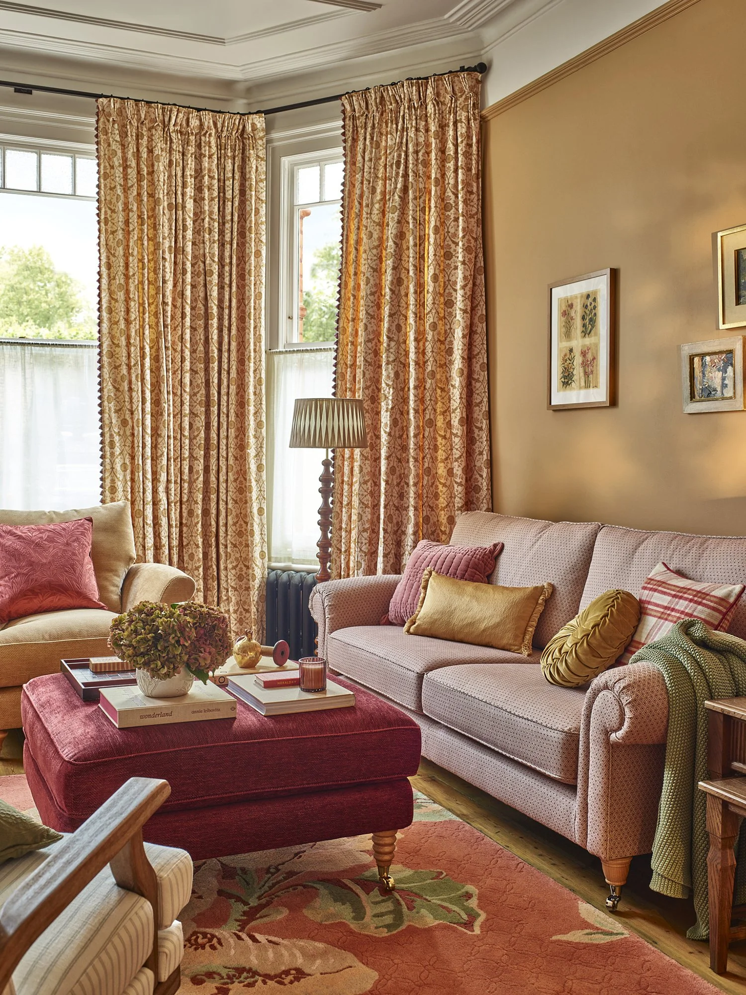 A cozy living room with patterned pink and beige curtains, pink and beige sofas with colorful pillows, a wooden side table, a flower arrangement, and a red upholstered ottoman on a floral area rug.