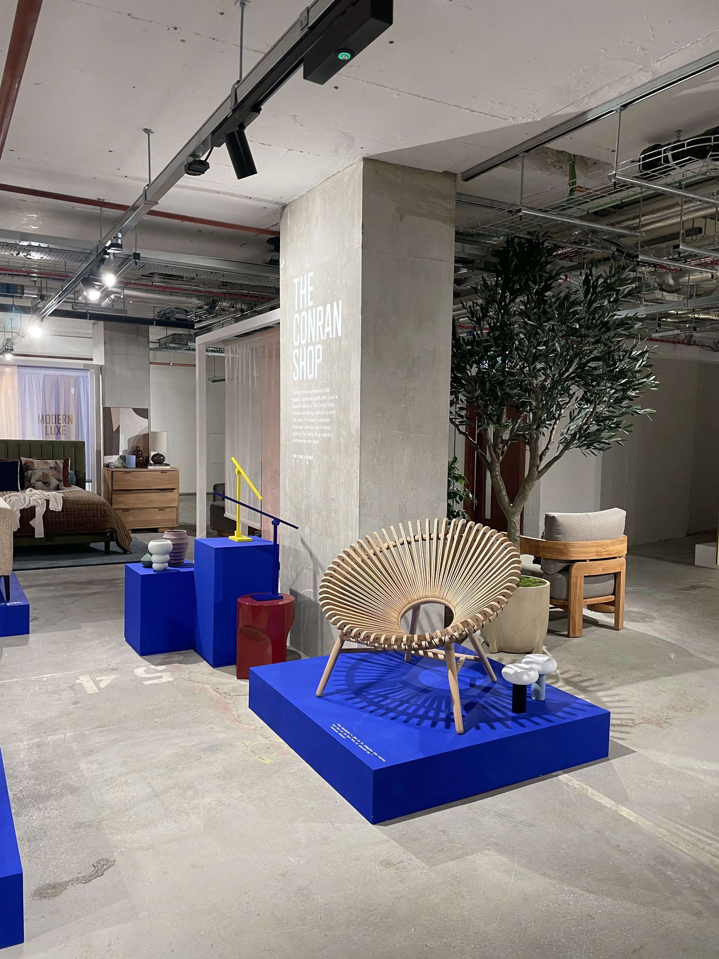 Interior of a showroom with modern furniture and decor items on display, including a rattan chair on a blue platform, a large potted plant, and smaller decorative objects.