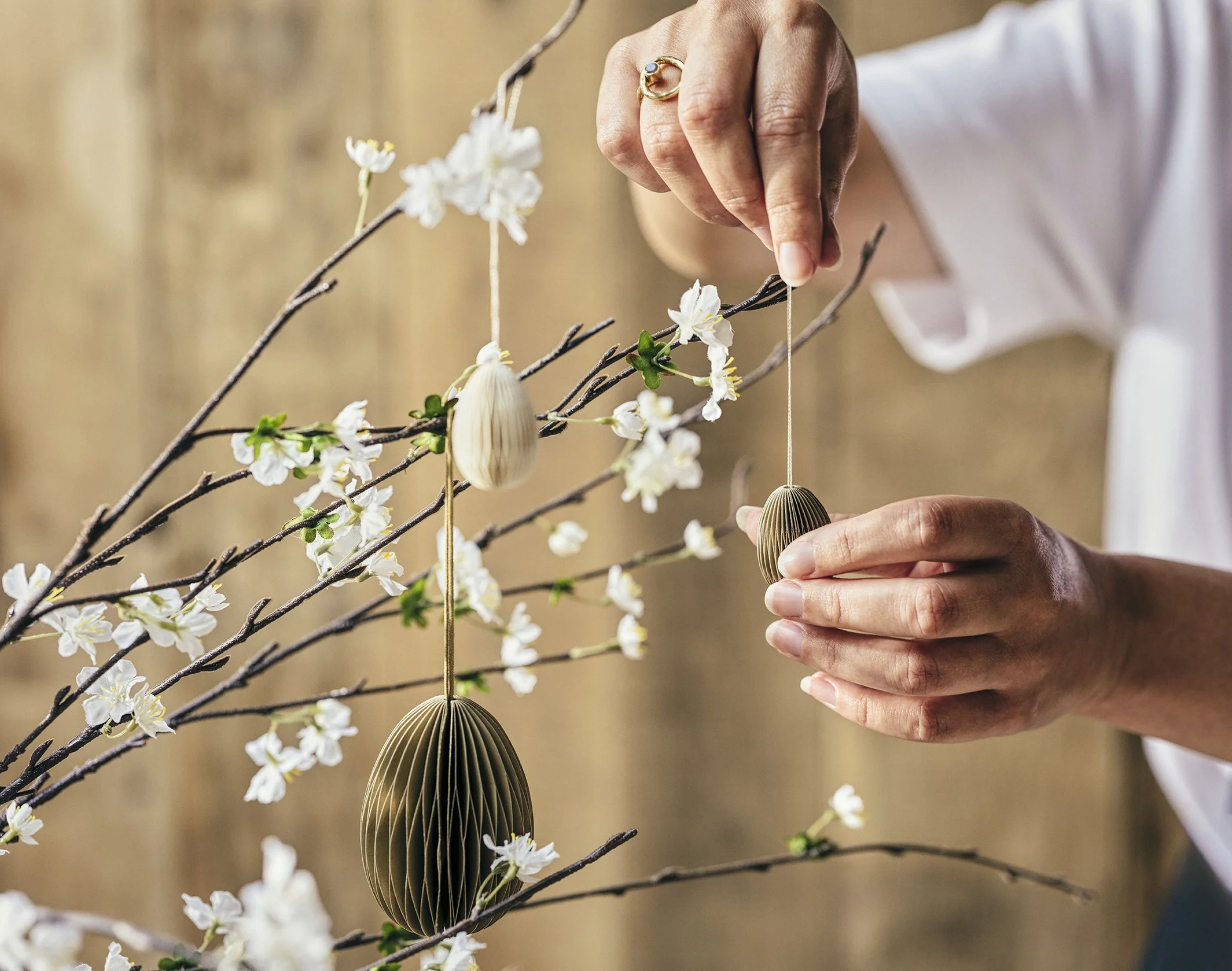 A person is hanging decorative egg ornaments on a flowering branch, likely for Easter, with a blurred background.