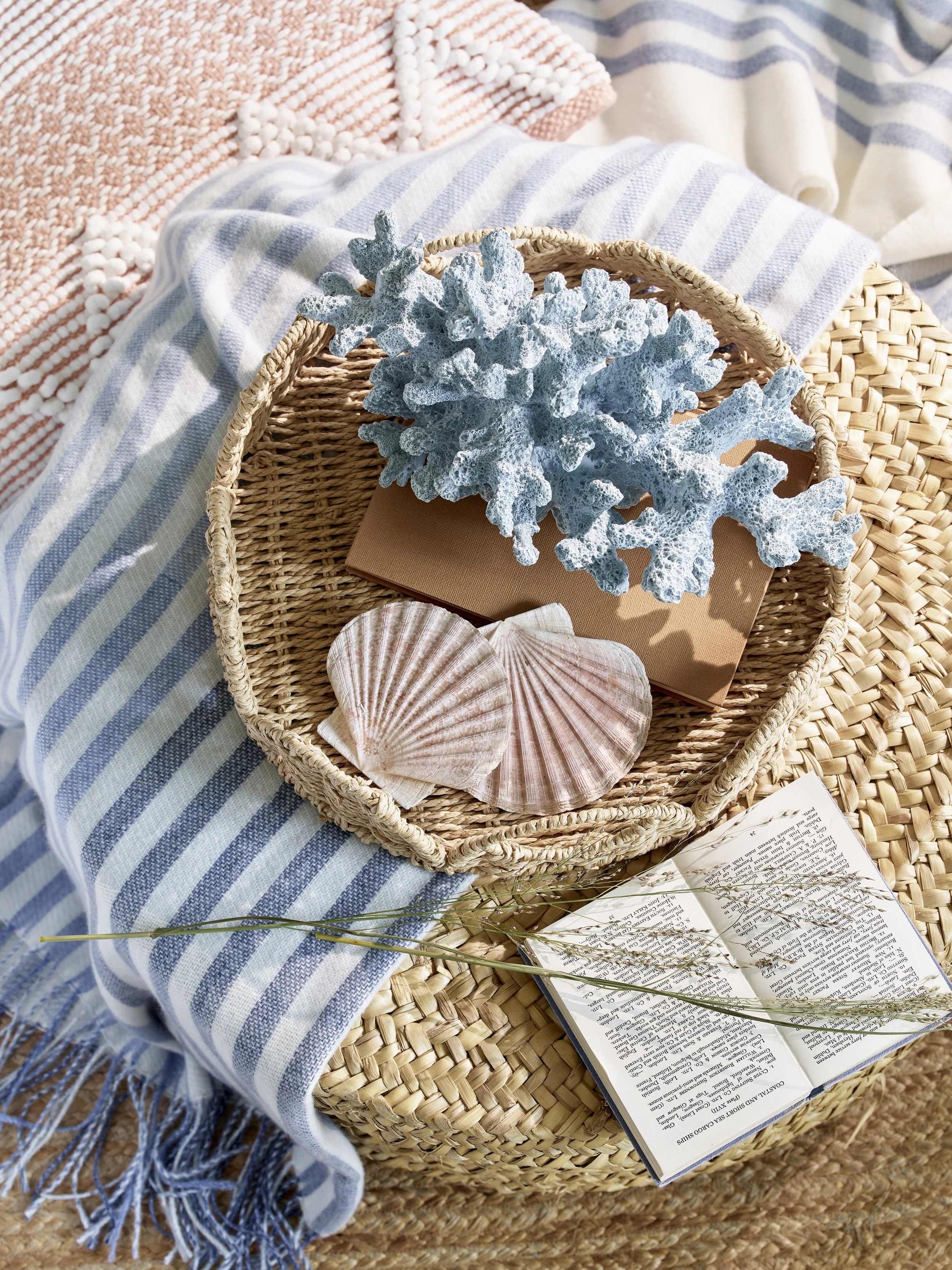 A woven basket containing a decorative blue coral branch, two seashells, and a brown notebook, placed on a striped blue and white blanket with a fringed edge, beside an open book with a branch on it, on a round woven mat.