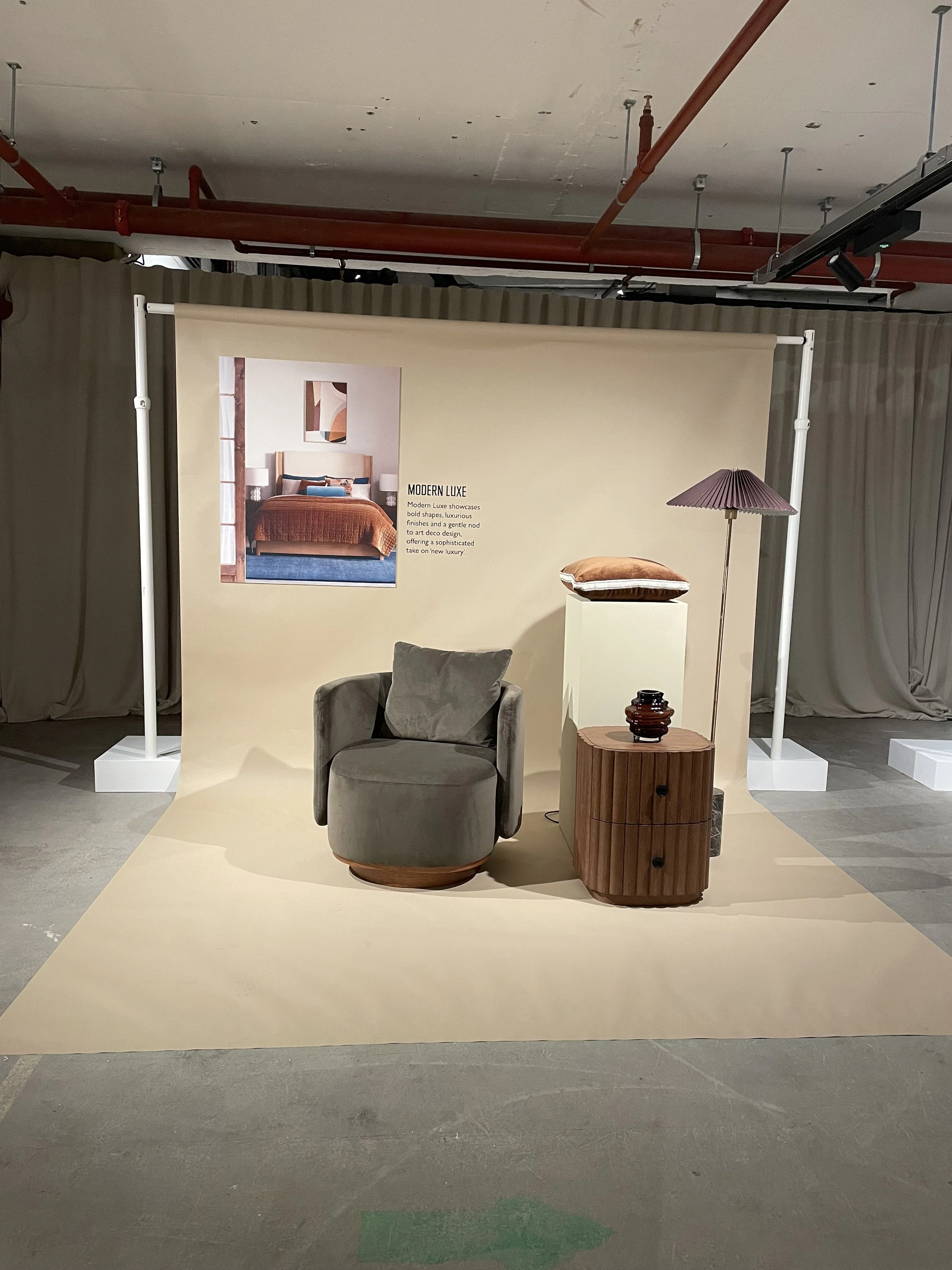 Display setup for modern rustic interior furniture with a gray armchair, a wooden nightstand with three drawers, a decorative table lamp with a purple shade, and a folded blanket or pillow on a white pedestal. A beige backdrop and a small poster show