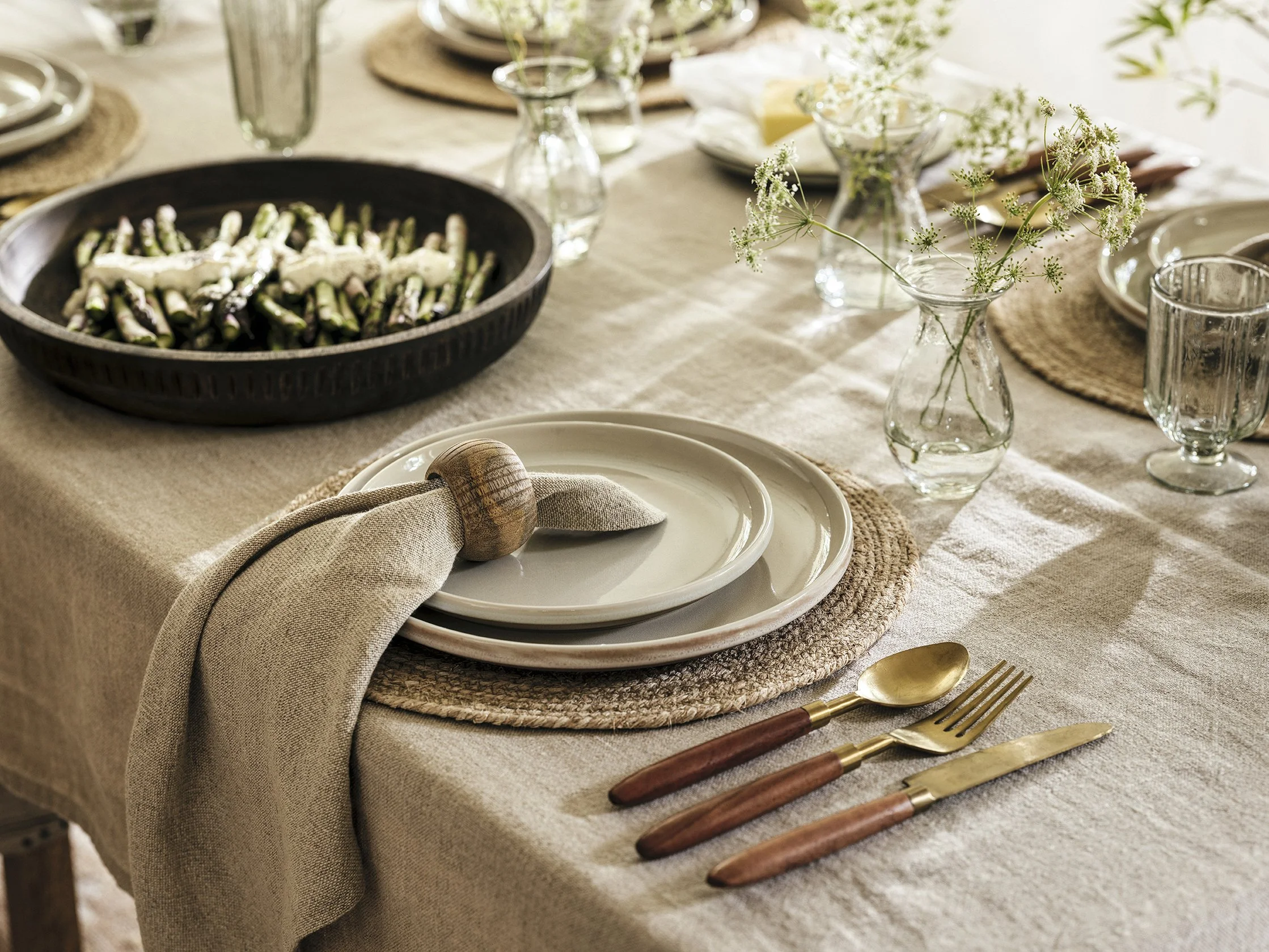A cozy dining table decorated with natural and neutral tones, featuring a beige tablecloth, woven placemats, arranged plates, gold-toned cutlery with wooden handles, clear glass vases with white flowers, a black serving bowl of asparagus with a cream