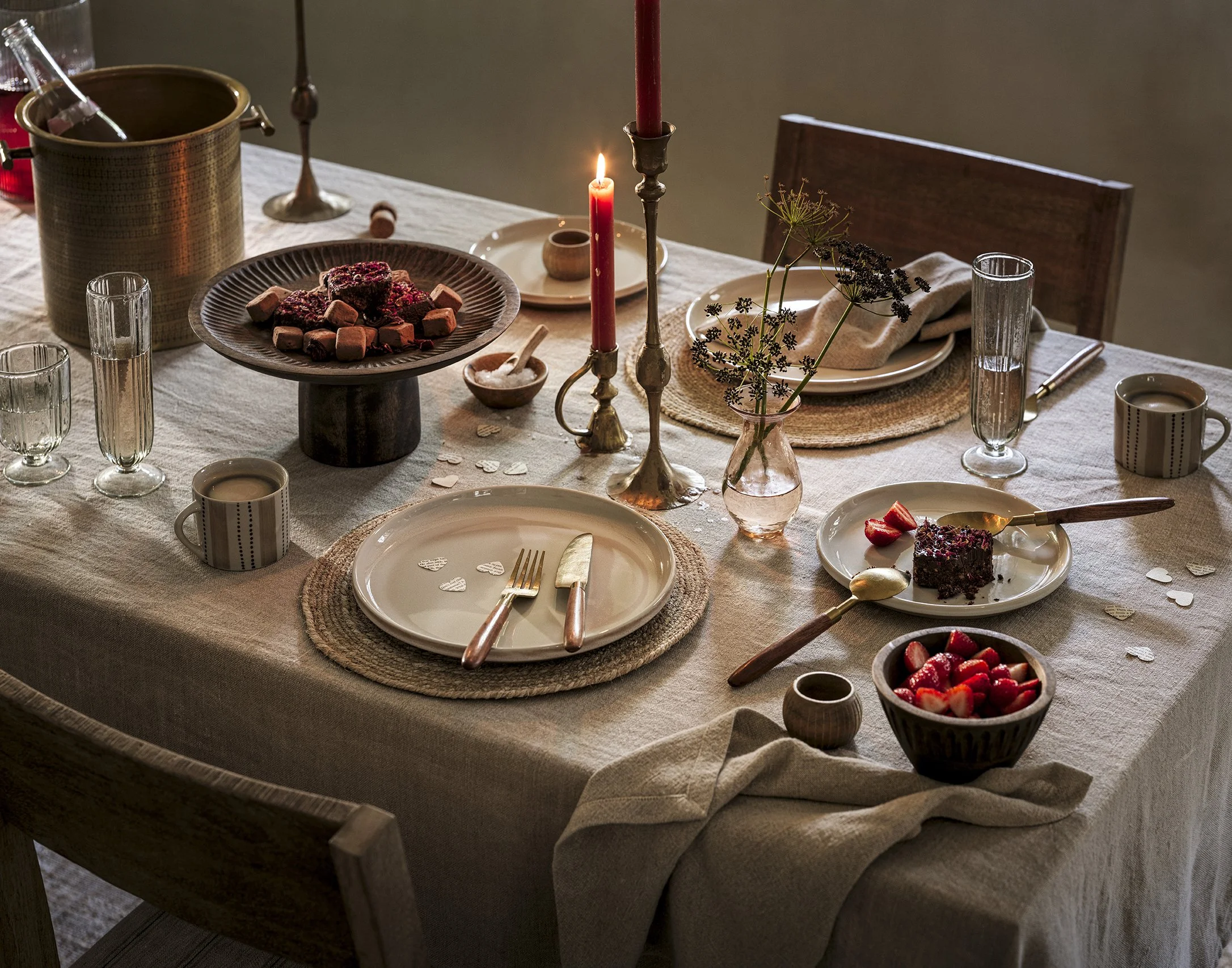 A table set for a meal with plates, glasses, a bowl of strawberries, a cake with berries, candles, and a vase with flowers.