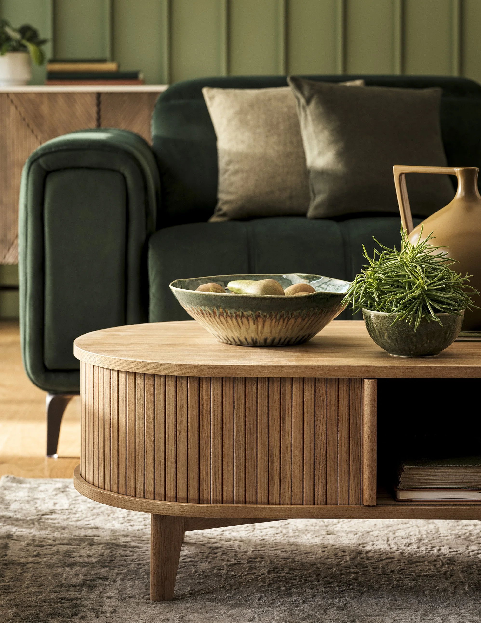 Living room with a green velvet sofa, beige pillows, a wooden coffee table with a bowl of stones, a potted plant, and a beige vase.