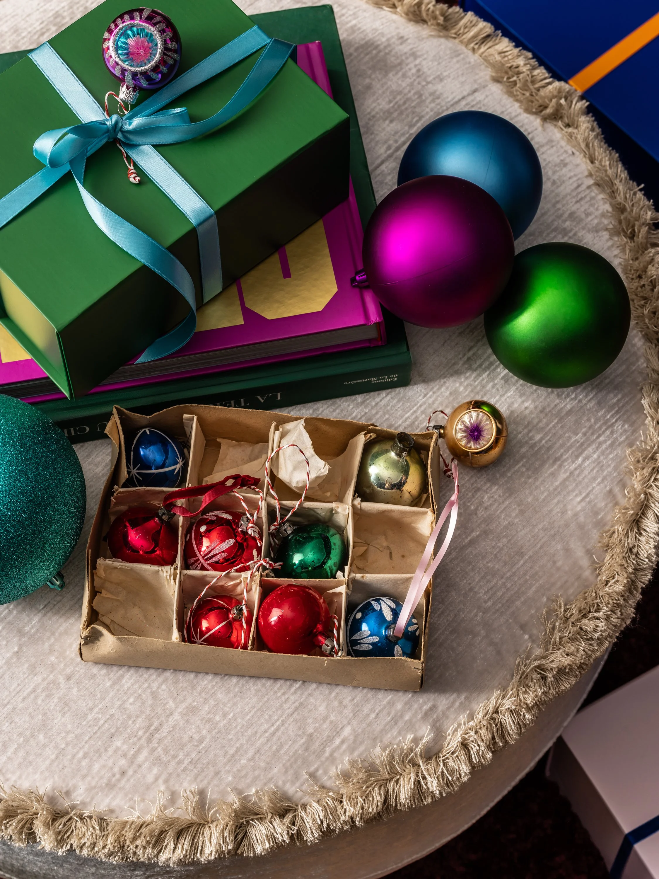 A table with Christmas ornaments, including a box of glass baubles in red, gold, green, blue, and silver, some with ribbons, next to wrapped gifts and colored balls in purple, blue, green, and teal.