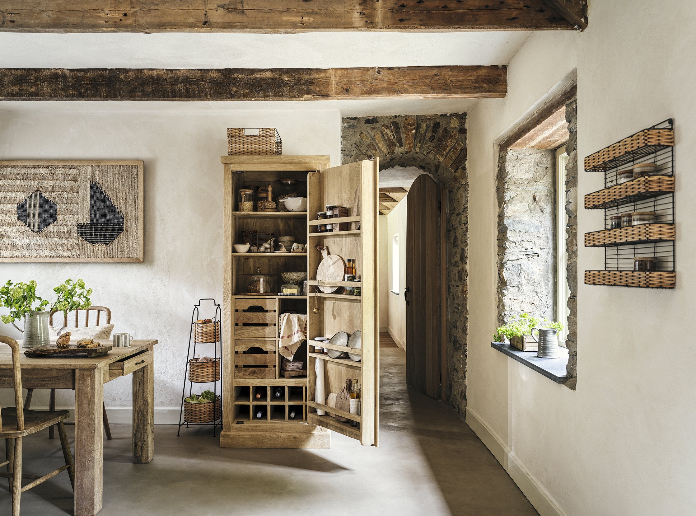 A rustic kitchen with wooden furniture, open cabinets, and stone walls, featuring a dining table with a pitcher and two glasses, and a window with potted plants.
