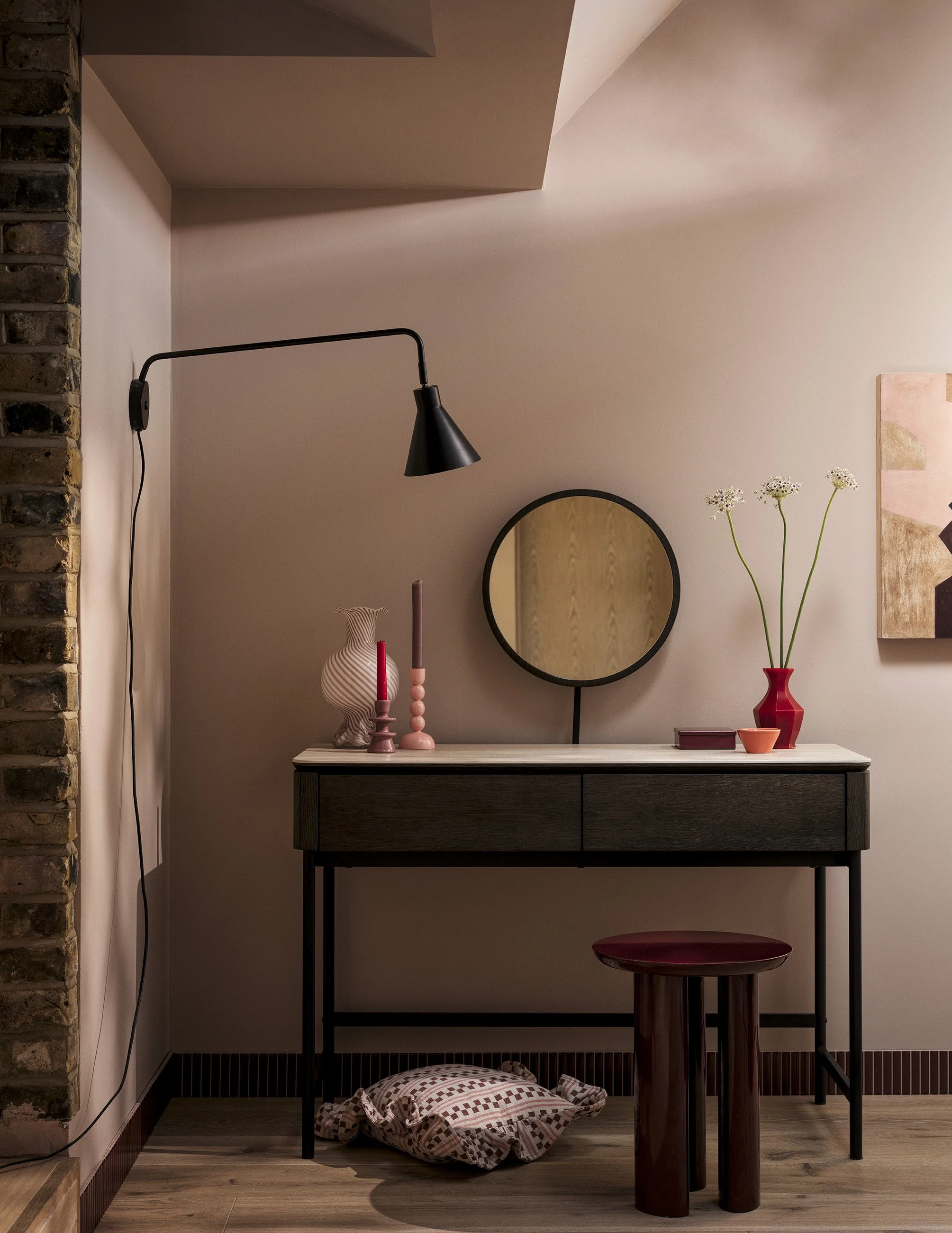 A decorated hallway corner with a dark wood and white marble console table, a round wall mirror, a red vase with tall white flowers, pink and red candles, a small closed box, a cushion on the floor, a black wall-mounted lamp, exposed brick wall on th