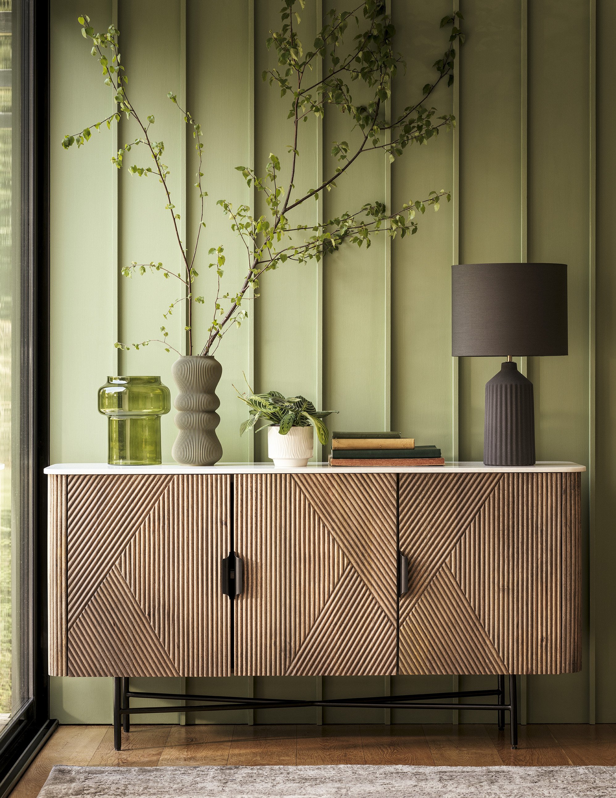 A modern wooden sideboard with vertical ribbed design against a green paneled wall. Decor includes a tall branch in a textured gray vase, a small green glass jar, a potted plant, a black table lamp, and stacked books.