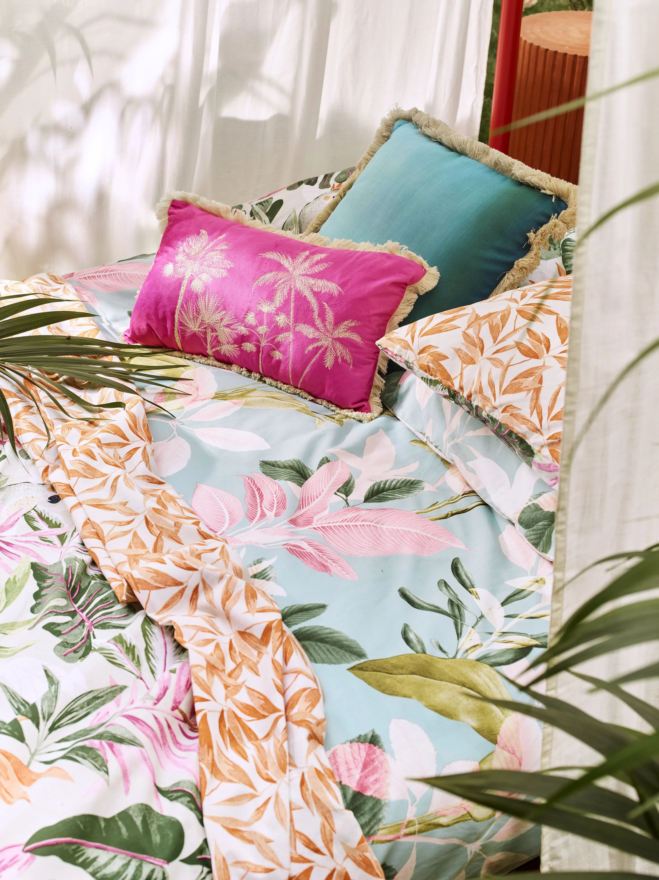 Colorful bed with tropical-themed bedding, including pink, orange, and green leaves, and decorative pillows in pink, blue, and patterned orange and cream.