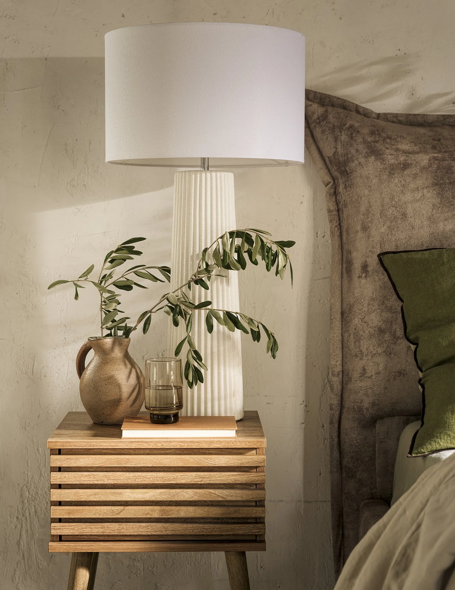 A bedside table with a ceramic vase holding a green leafy branch, a glass of dark liquid, and a closed book, topped with a tall white textured lamp, next to a bed with a beige headboard and a green pillow.