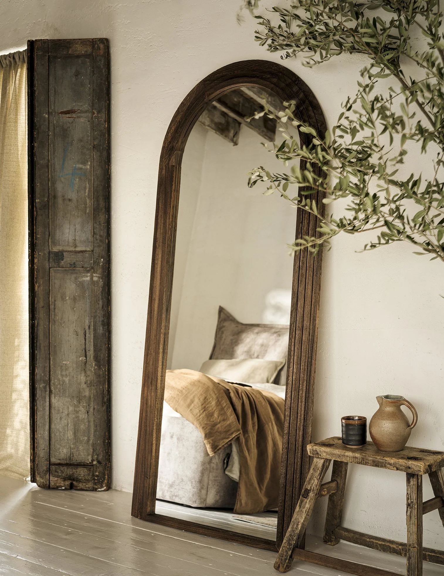A rustic bedroom with a wooden-framed mirror leaning against a white wall, a small wooden stool with a pitcher and a cup, and a partial view of a bed with beige and brown bedding, a pillow, and a blanket.