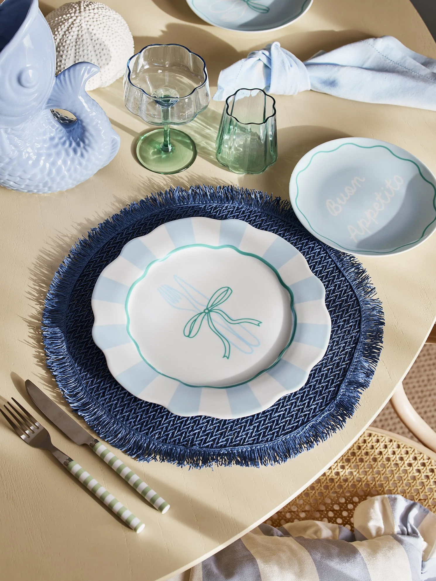 A table setting with a blue woven placemat, a decorative plate with a fork and knife with striped handles, and two ceramic bowls, one with the words 'Buen Apetito'. There are also two glass cups, a white ceramic fish-shaped teapot, a white textured b