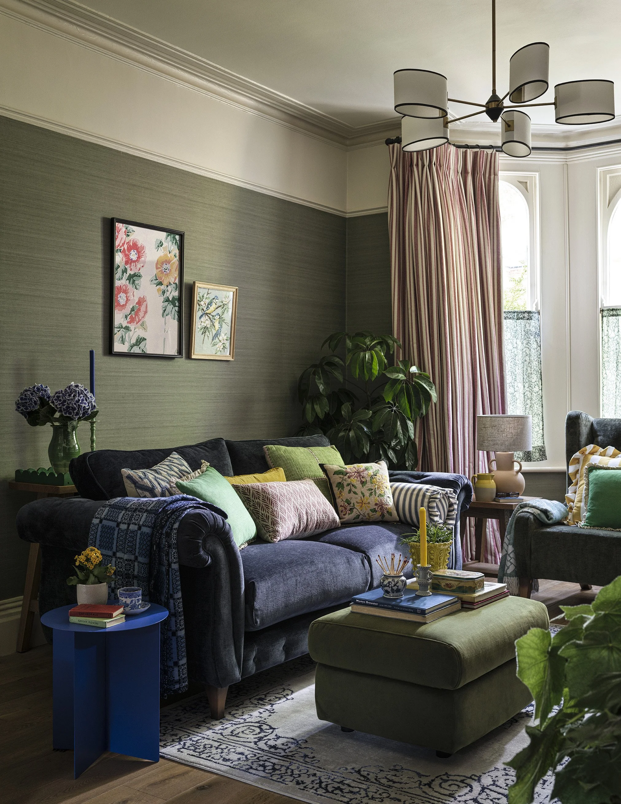 Living room with navy sofa, green ottoman, colorful cushions, framed wall art, large window with red striped curtains, table lamp, indoor plants, and decorative items.