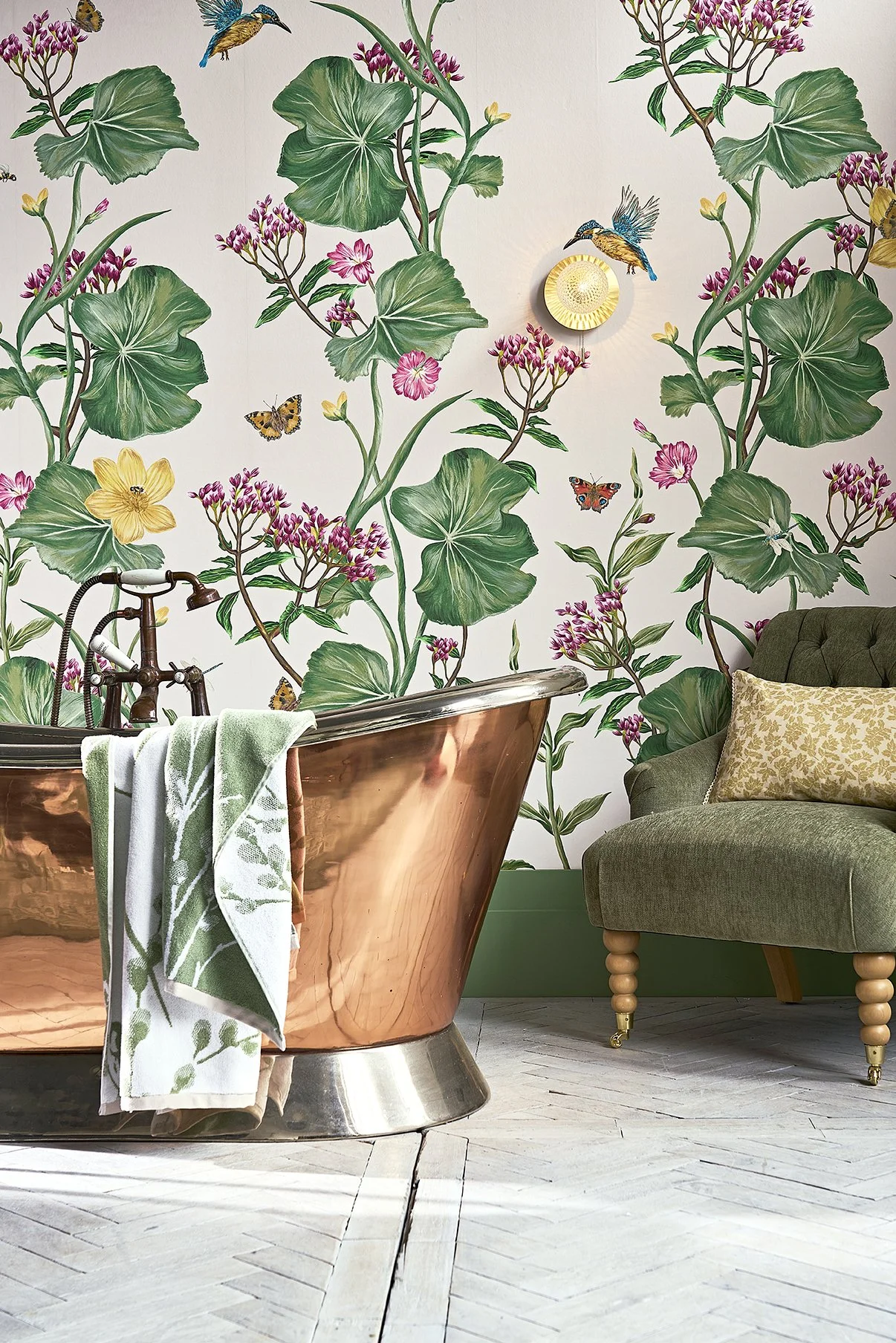 A copper bathtub with a green and white towel draped over the side, a vintage green armchair with a gold patterned pillow, a floral and butterfly wallpaper, and a wall-mounted light fixture.