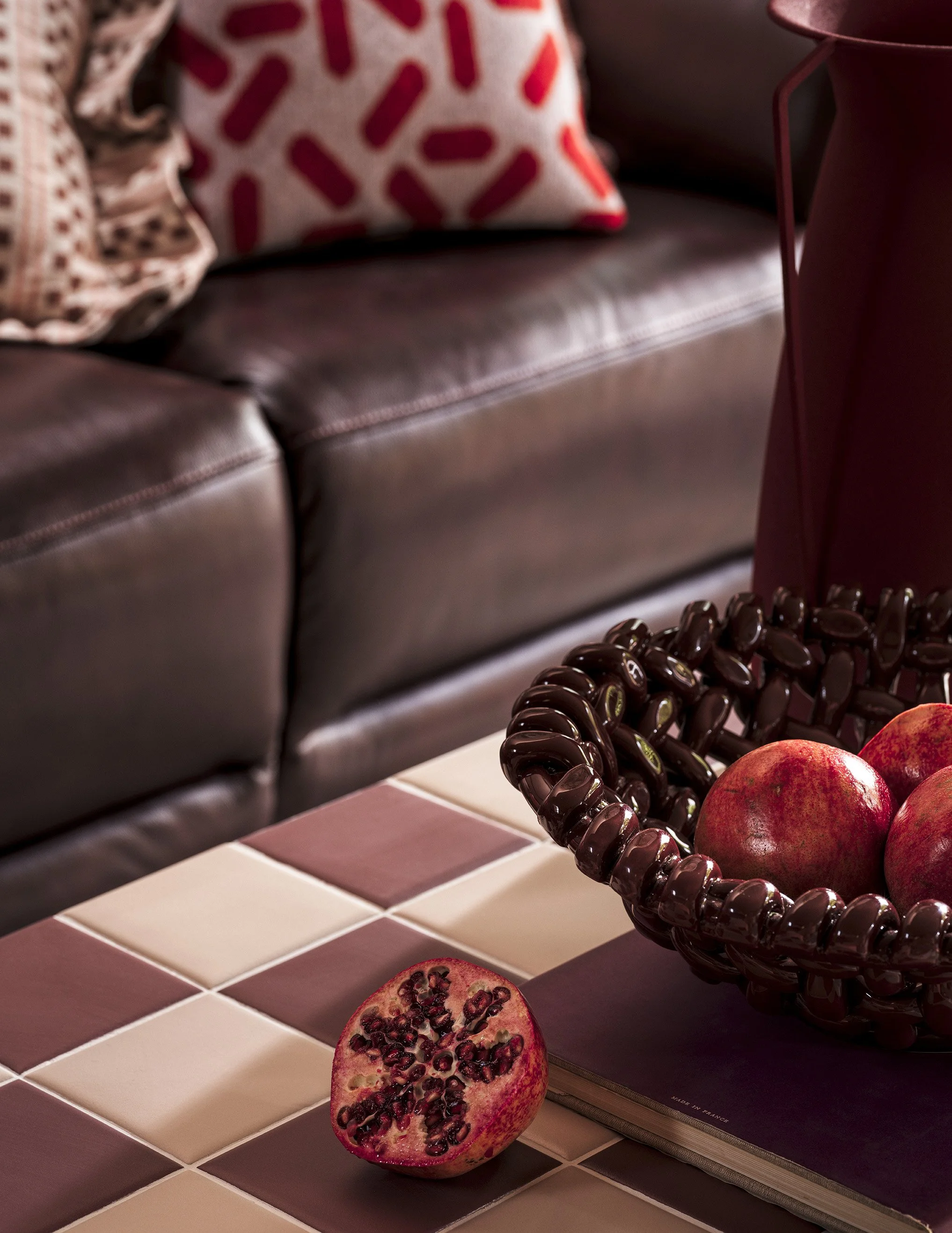 Close-up of a checkered tile table with a halved pomegranate, a basket of whole pomegranates, and a black book. A leather sofa with decorative pillows is in the background.
