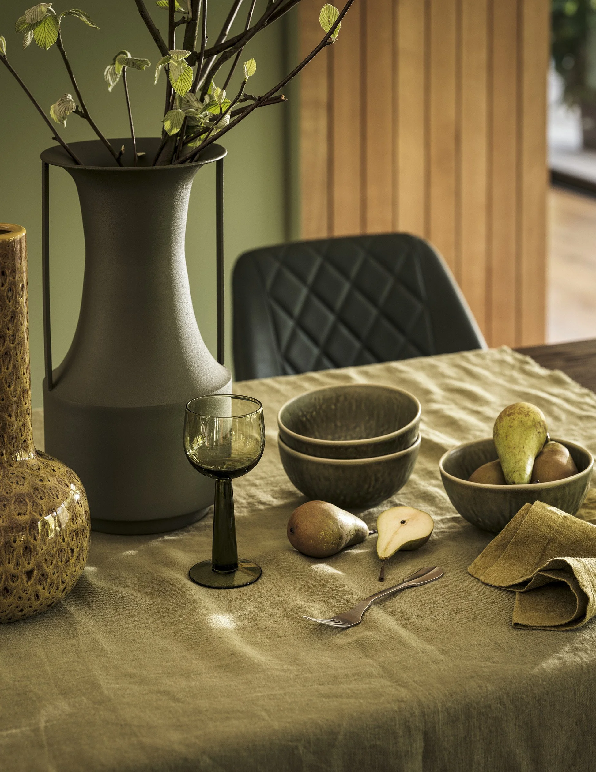 A dining table with a beige tablecloth, a green vase with branches, a wine glass, ceramic bowls, a pear, an apple, a fork, and a folded cloth napkin inside a room with a wooden wall and a black chair.