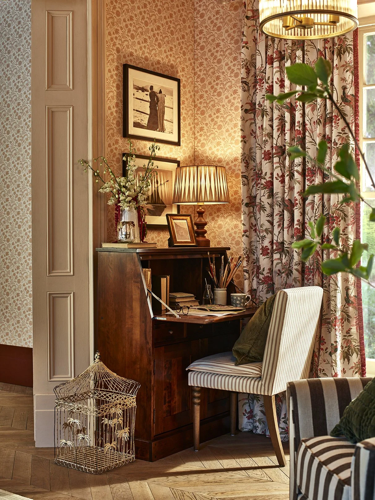 Cozy corner of a room with a wooden desk, striped chair, floral curtains, and a table lamp, decorated with framed photos, plants, and books.