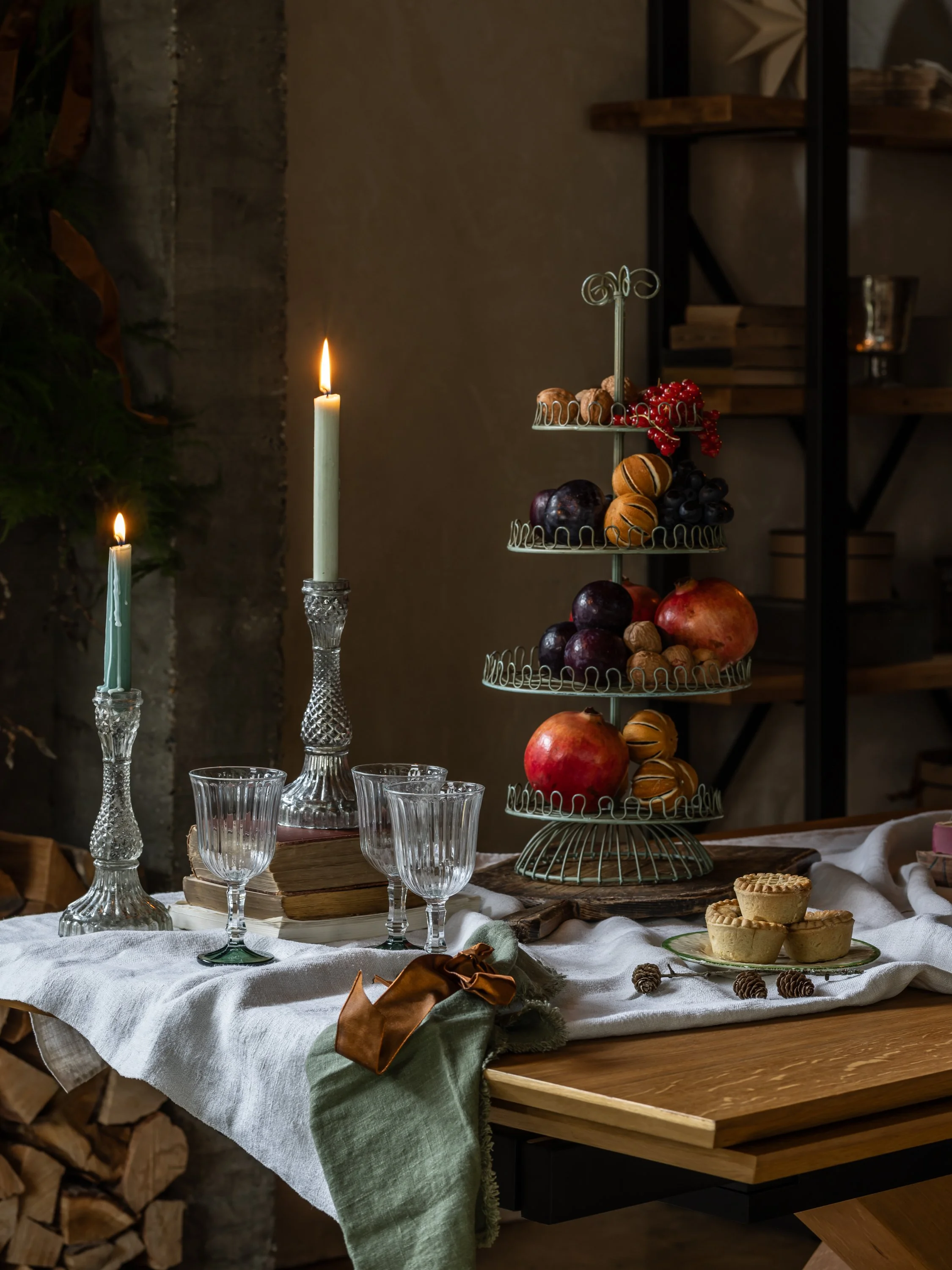 A rustic dining table decorated for a special occasion with two lit candles in crystal holders, empty crystal glasses, a stack of vintage books, a tiered fruit stand with apples, grapes, pomegranates, and assorted desserts, and pinecones scattered on